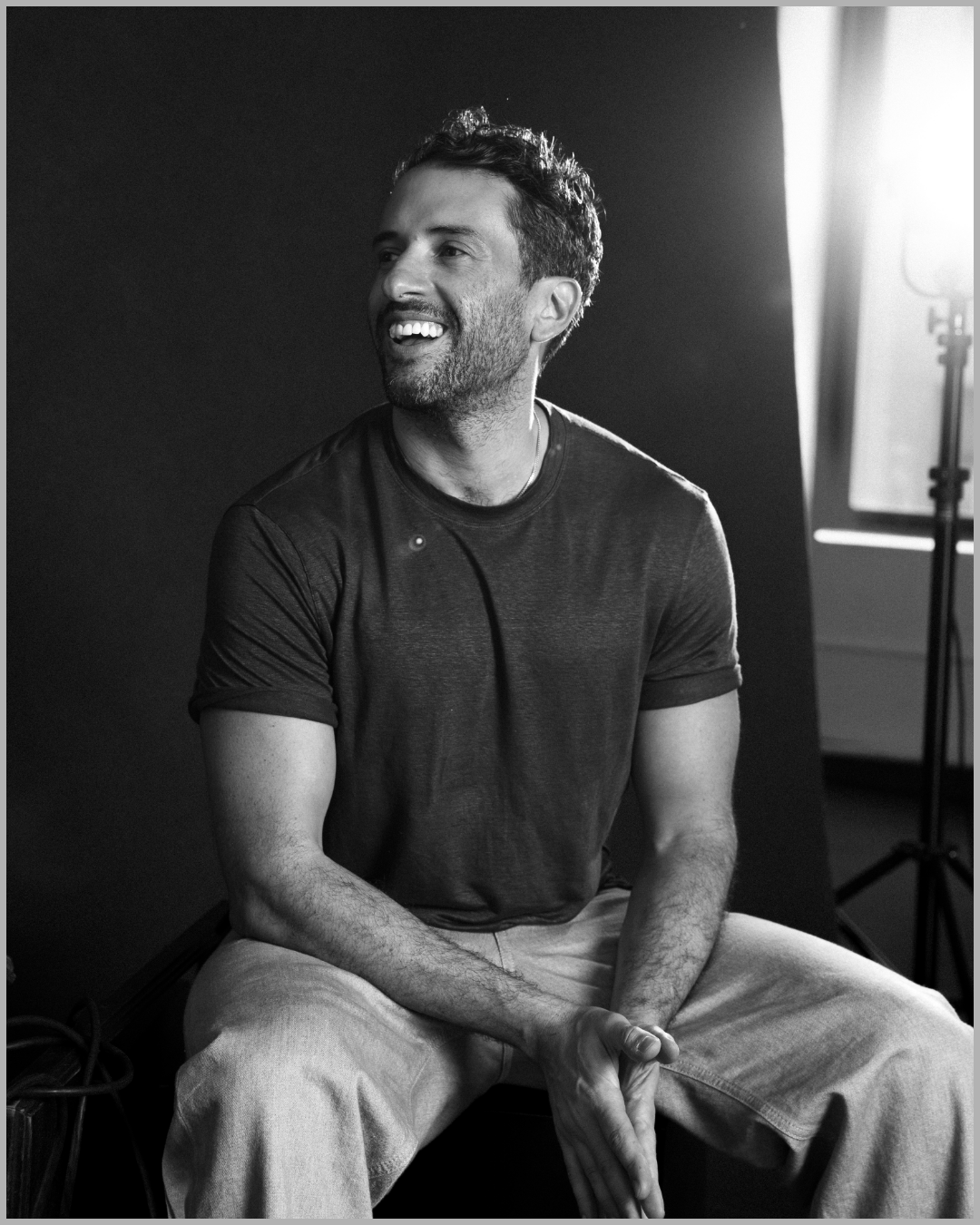 A smiling man with a beard and short curly hair, wearing a dark t-shirt and light-colored pants, sitting in front of a photography backdrop with studio lighting in the background.