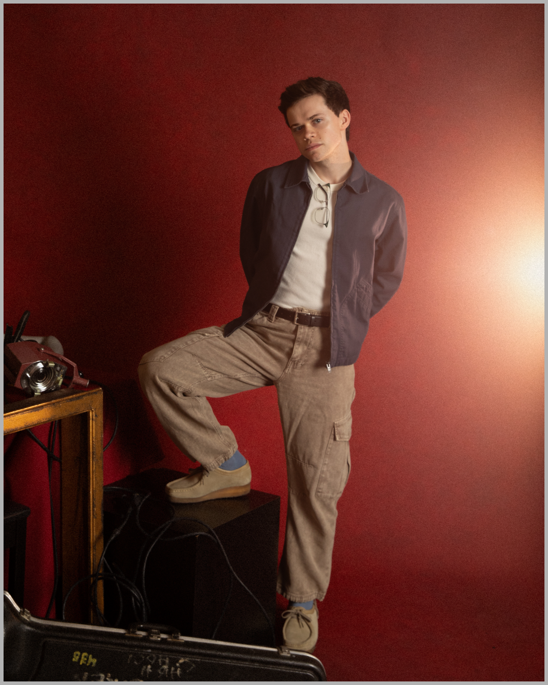 A young man with dark hair and fair skin standing against a red wall, wearing a dark jacket, beige cargo pants, and beige shoes, with glasses hanging from his shirt, with one foot resting on a black speaker and his hands behind his back.