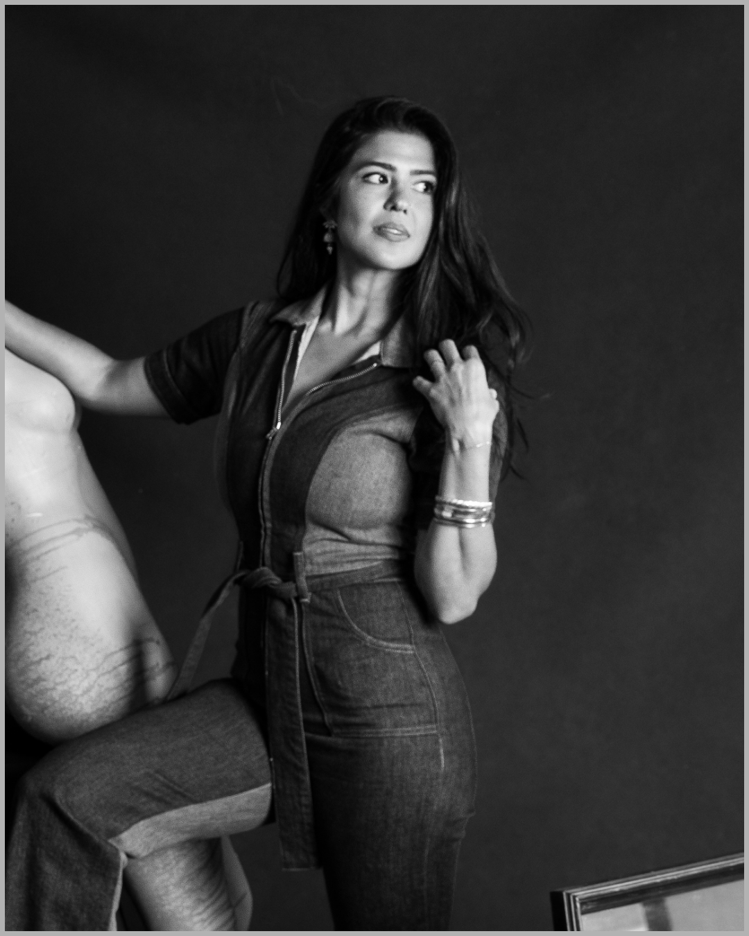 A woman with long dark hair, wearing a zip-up top and high-waisted jeans, standing against a plain dark background, looking to her right.