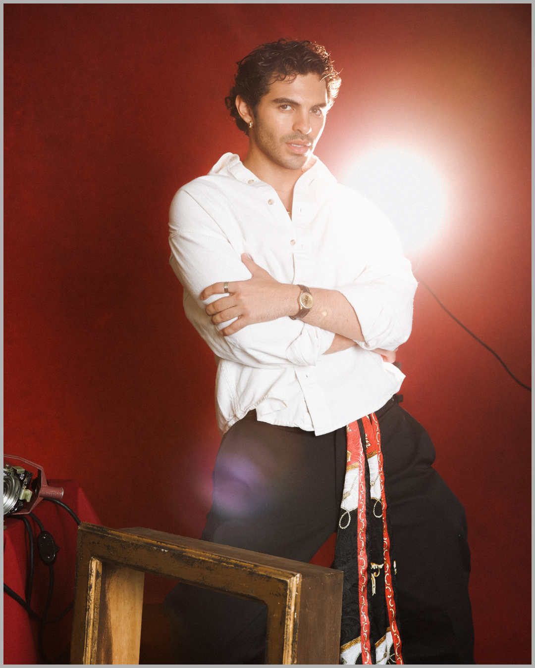 A man with curly dark hair, wearing a white shirt with rolled-up sleeves, black pants with a patterned scarf hanging from the waistband, and a gold watch, poses with crossed arms in front of a red background with a bright light behind him.