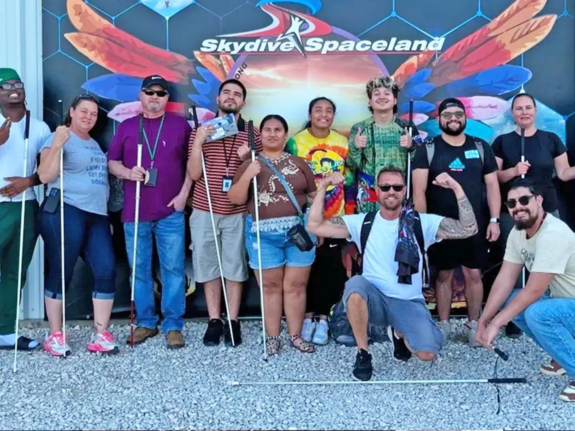 A group of smiling individuals stand in front of the Skydive Spaceland business in Austin, TX after facing the Visually Impaired and Blind Skydiving Challenge in 2024.