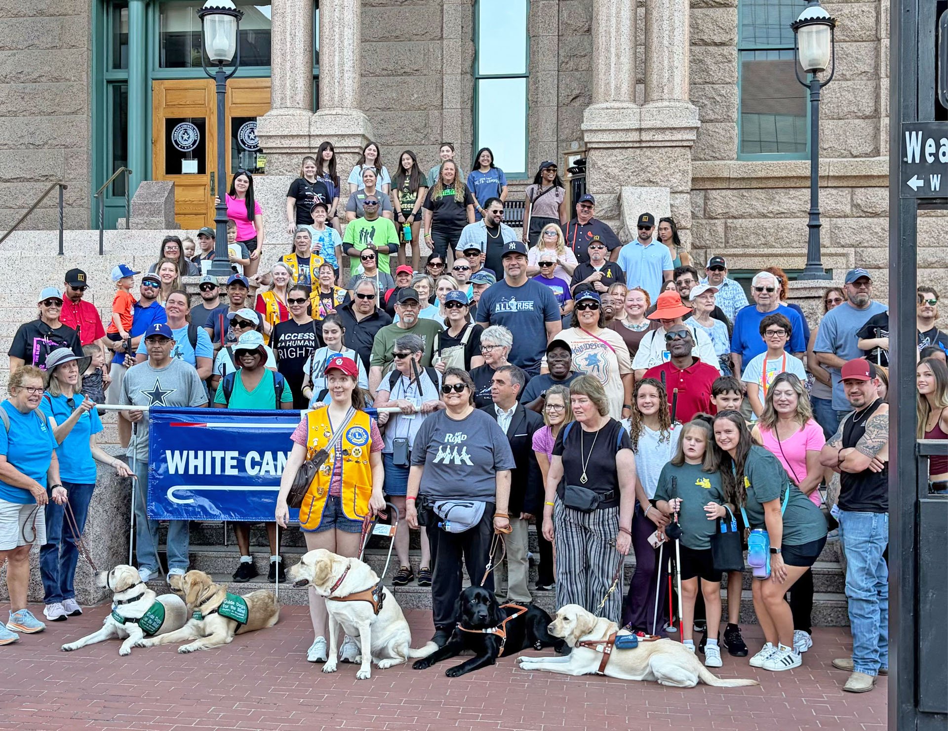 A large group of visually impaired and blind individuals stand on the Fort Worth County Courthouse steps, surrounded by seeing eye dogs and non-blind supporters after walking for White Cane Day.
