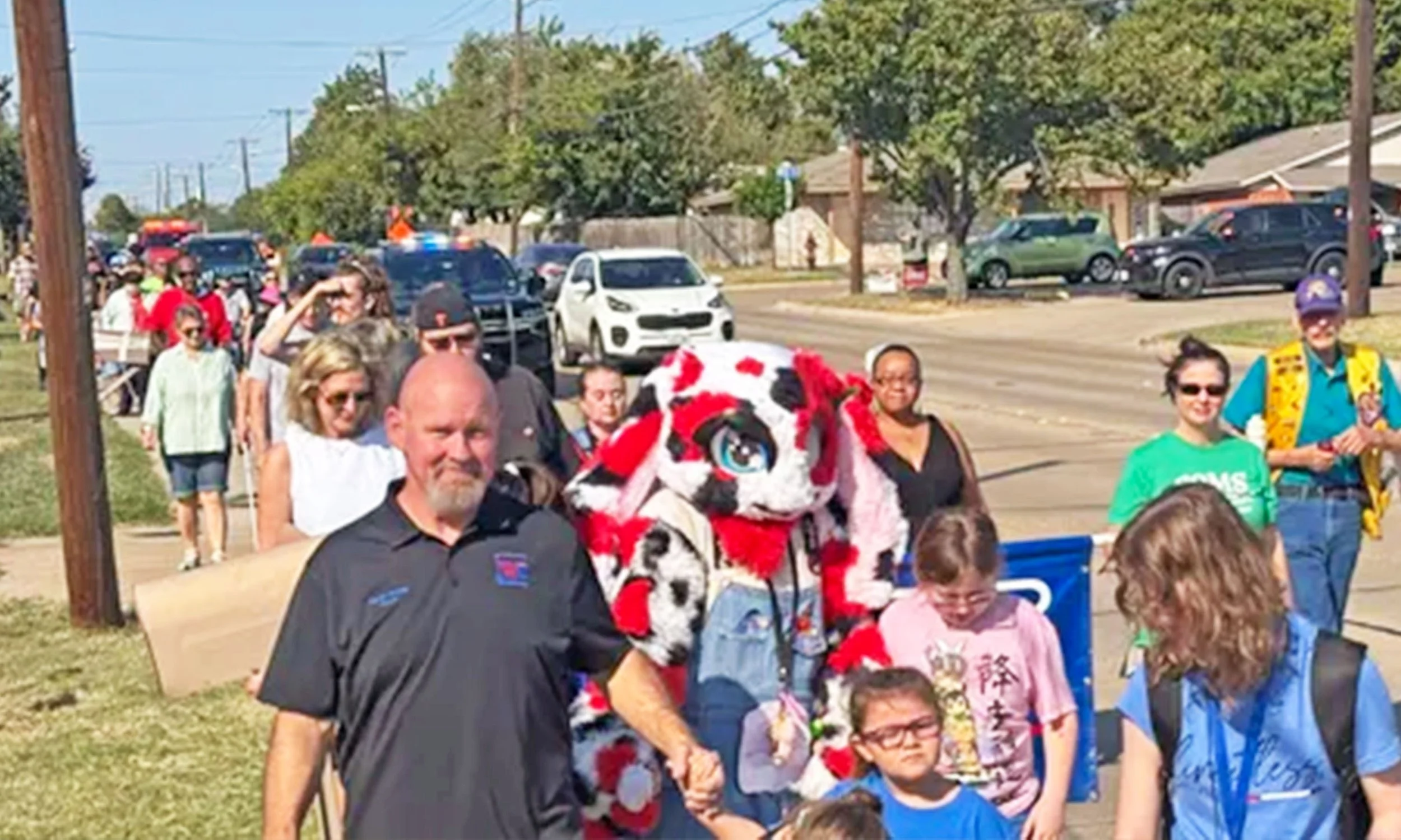 Mayor Faron Young of White Settlement leads a large group of supporters on foot through the town on White Cane Day 2024. Police cars escort the crowd with lights flashing.