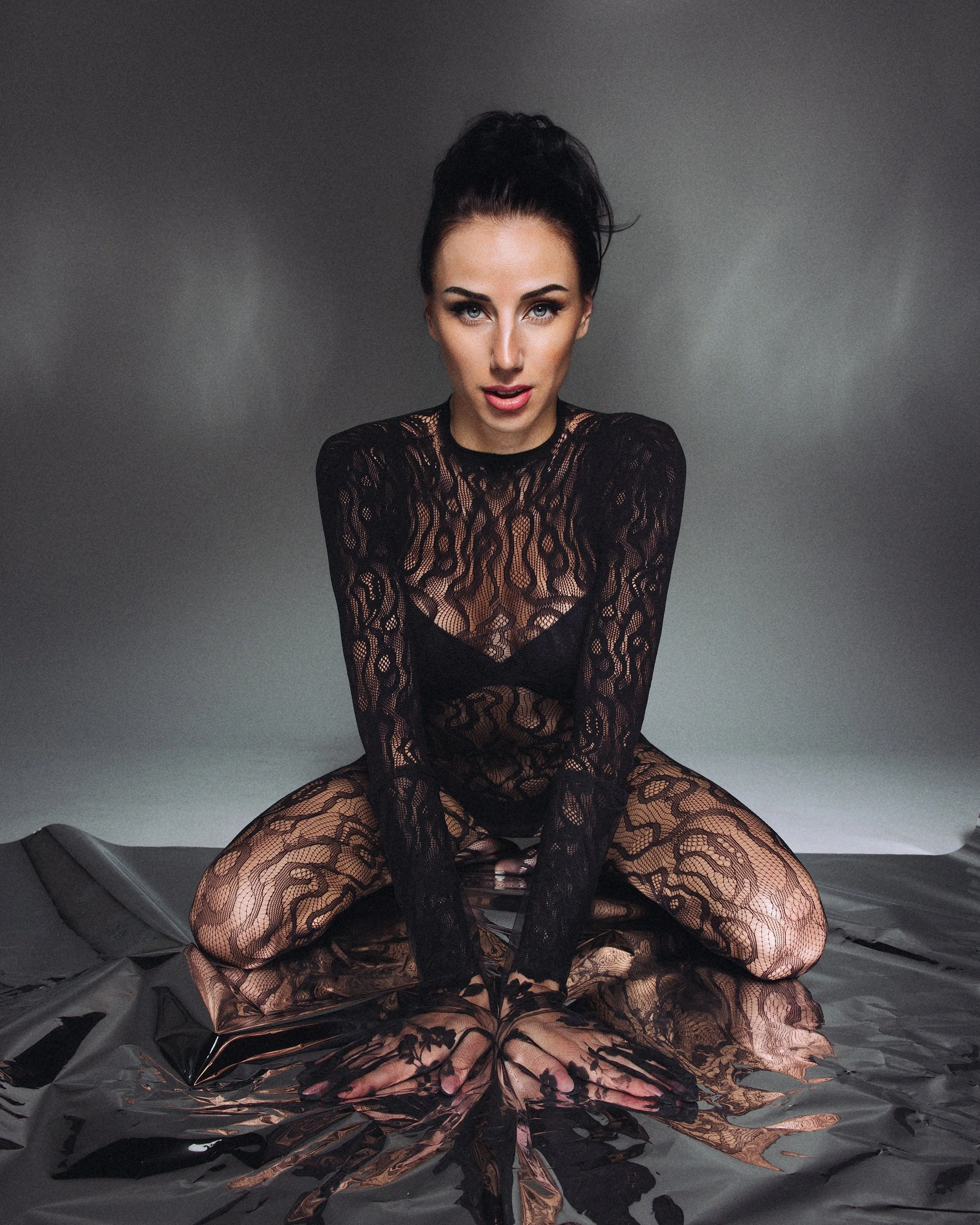 A woman with dark hair in a high bun wearing a black lace dress with long sleeves poses in front of a neutral gray background, sitting on crumpled silver foil.