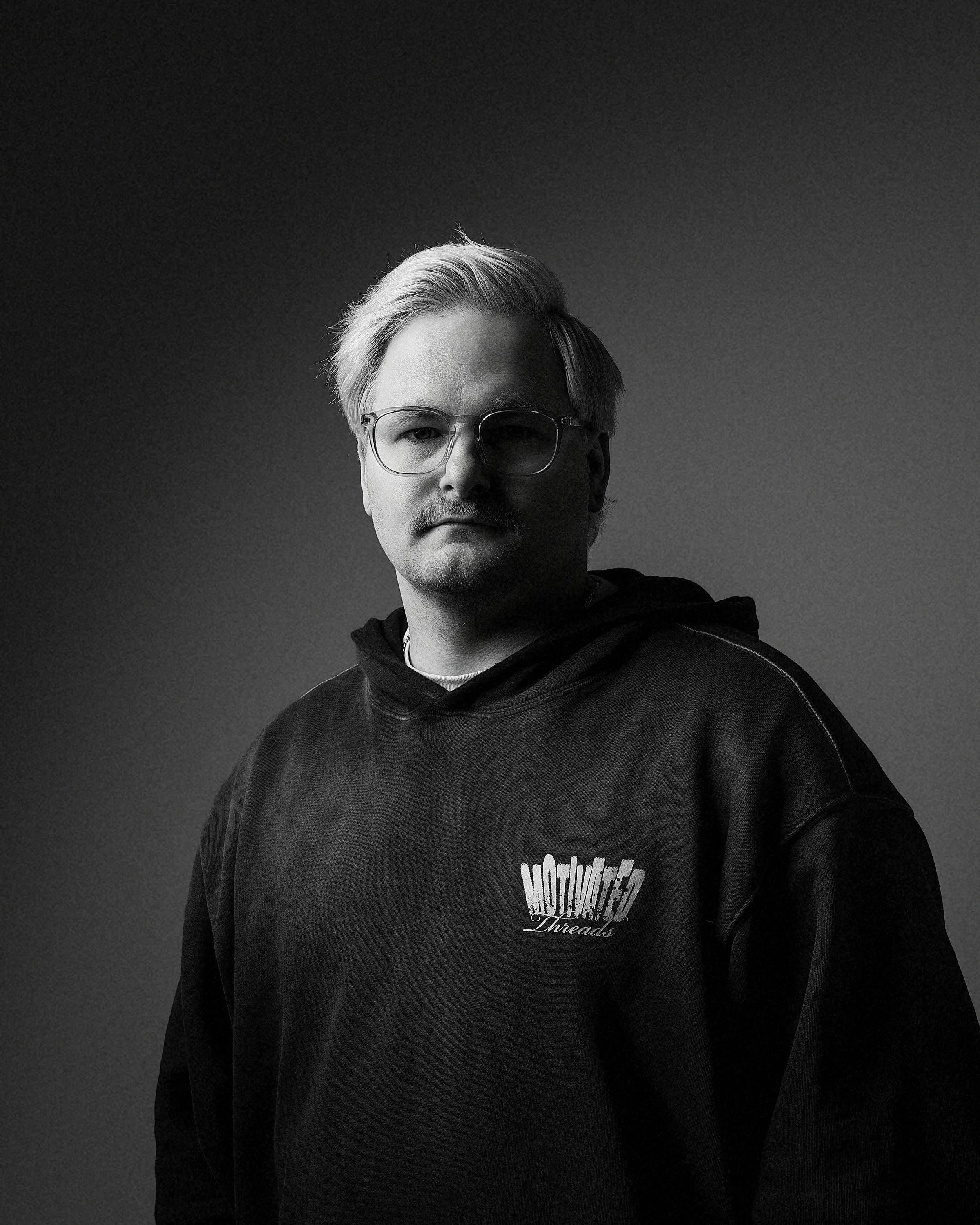 Black and white portrait of a man with glasses and blonde hair, wearing a hoodie with a 'Threads' logo, against a dark background.