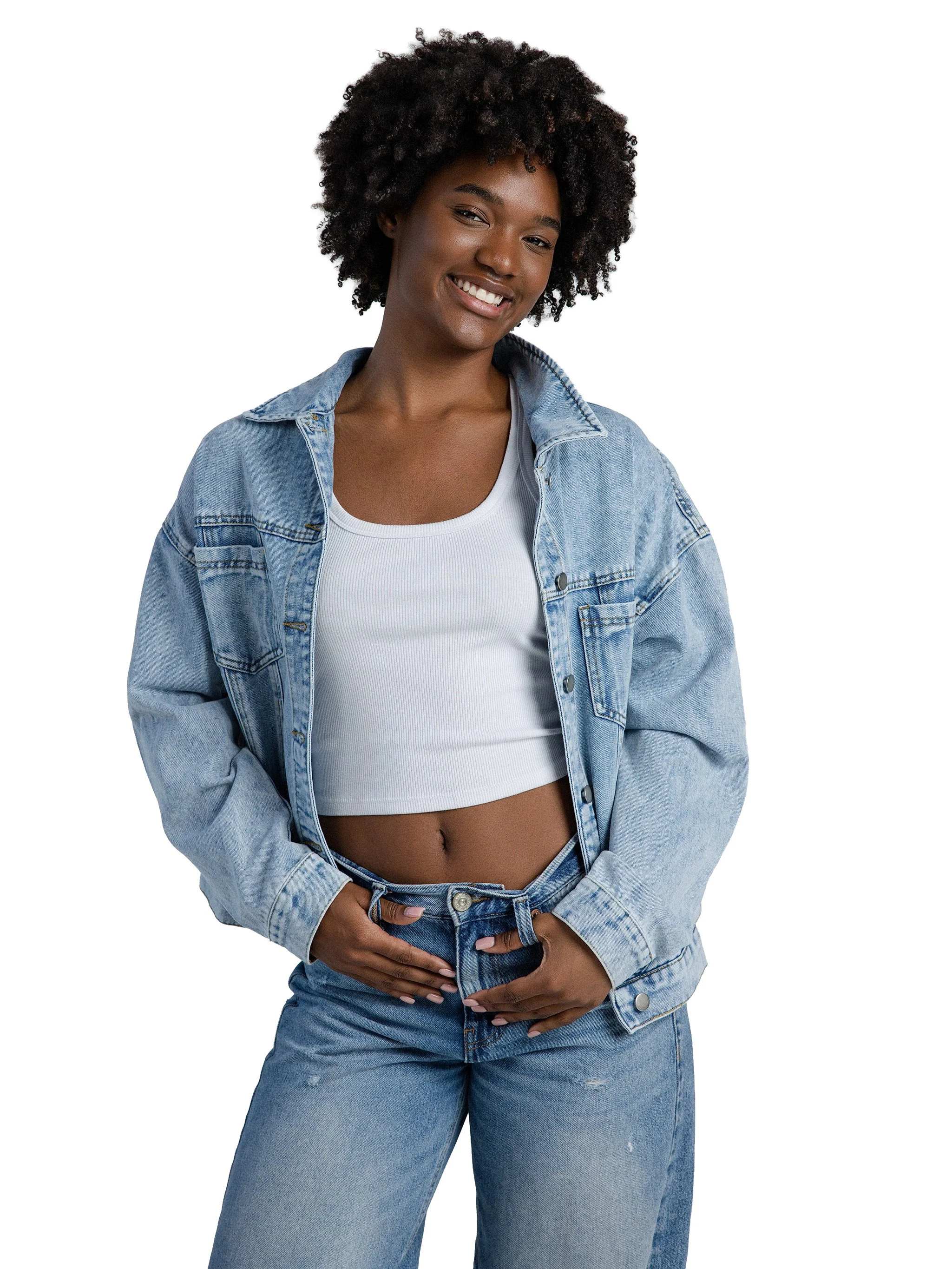 A woman with natural curly hair wearing a denim jacket, a white crop top, and jeans, smiling and positing with her hand on her stomach against a white background.