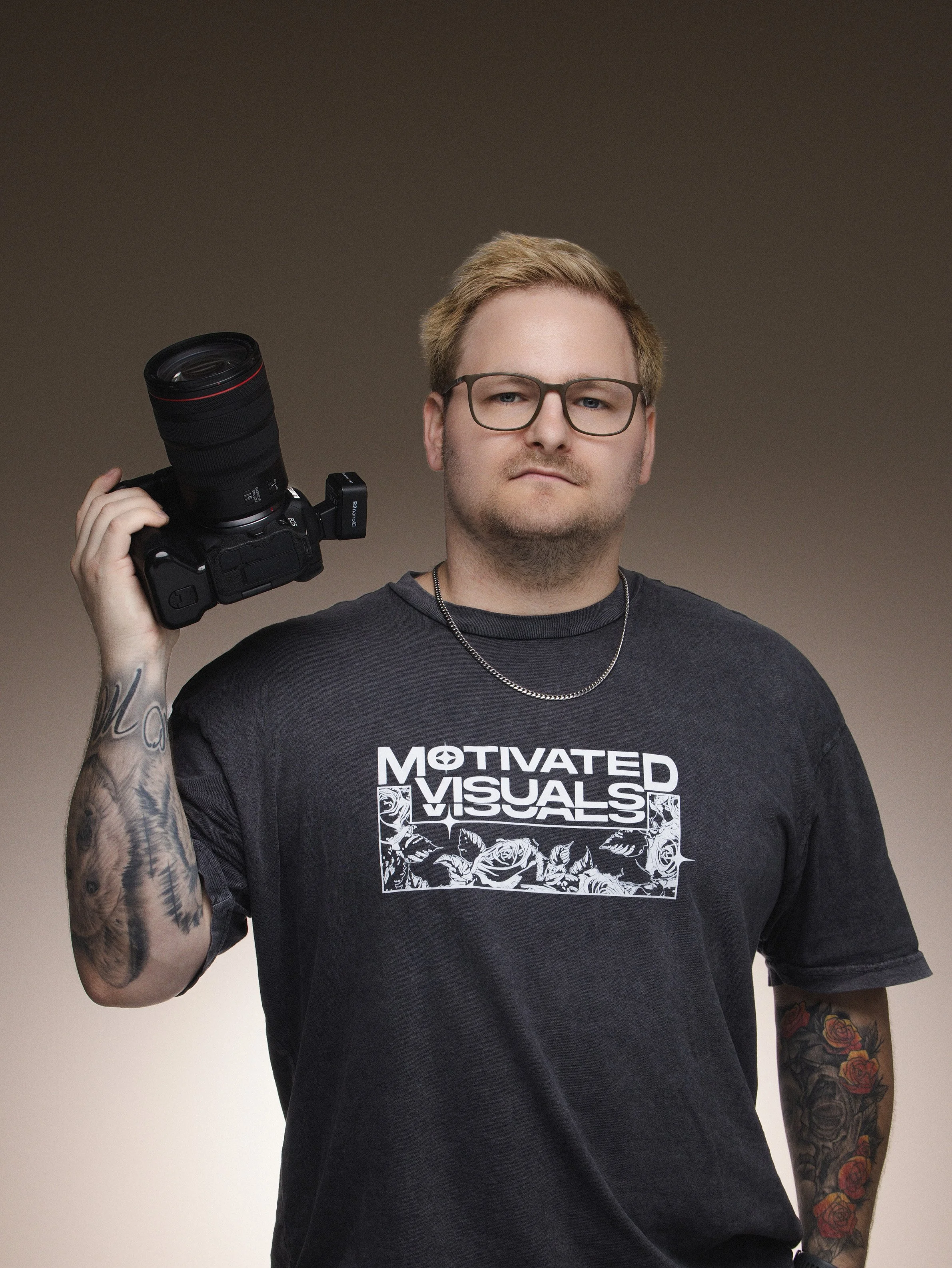 Young man with blond hair and glasses, holding a professional camera over his shoulder, wearing a black T-shirt that says 'Motivated Visuals', standing against a plain gradient background.