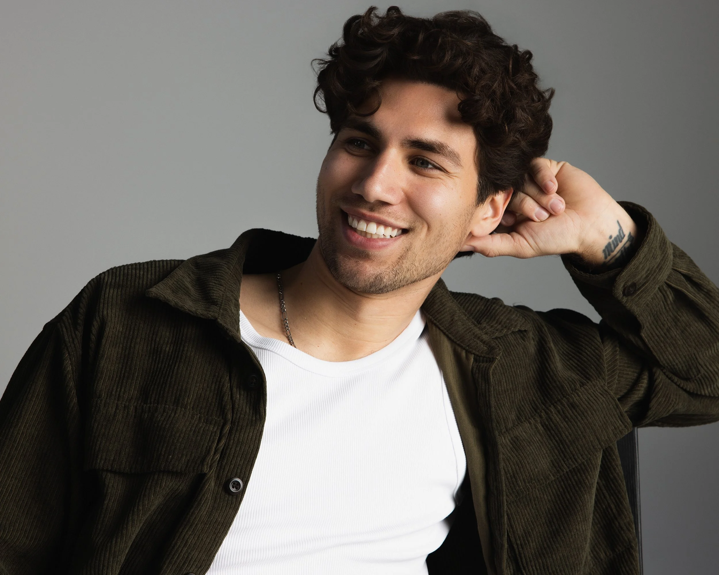 A young man with dark, curly hair smiling, wearing a black and olive green jacket over a white shirt, with a tattoo on his wrist, against a plain gray background.