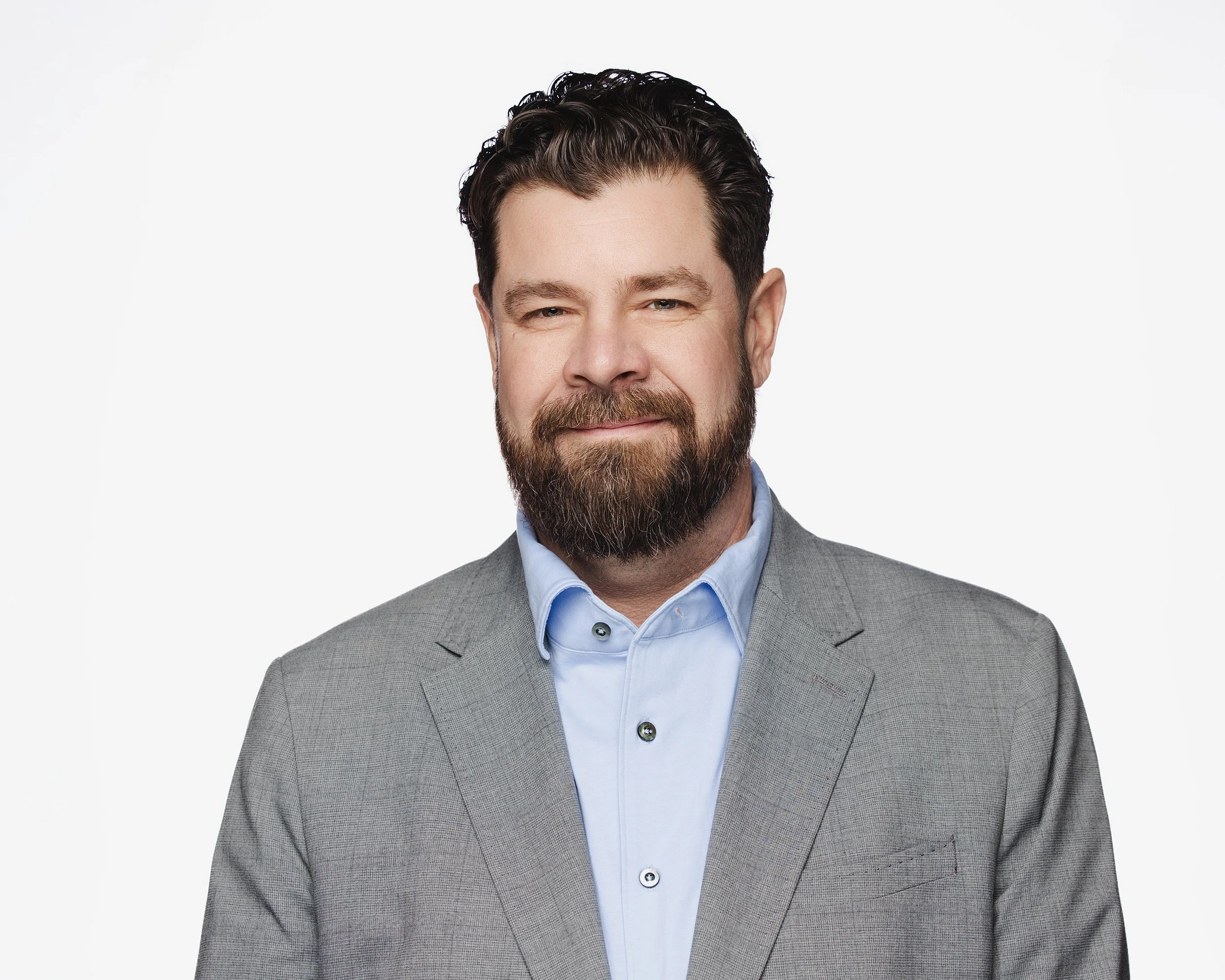 A man with a dark beard and hair, wearing a light blue dress shirt and a gray blazer, smiling slightly against a plain white background.
