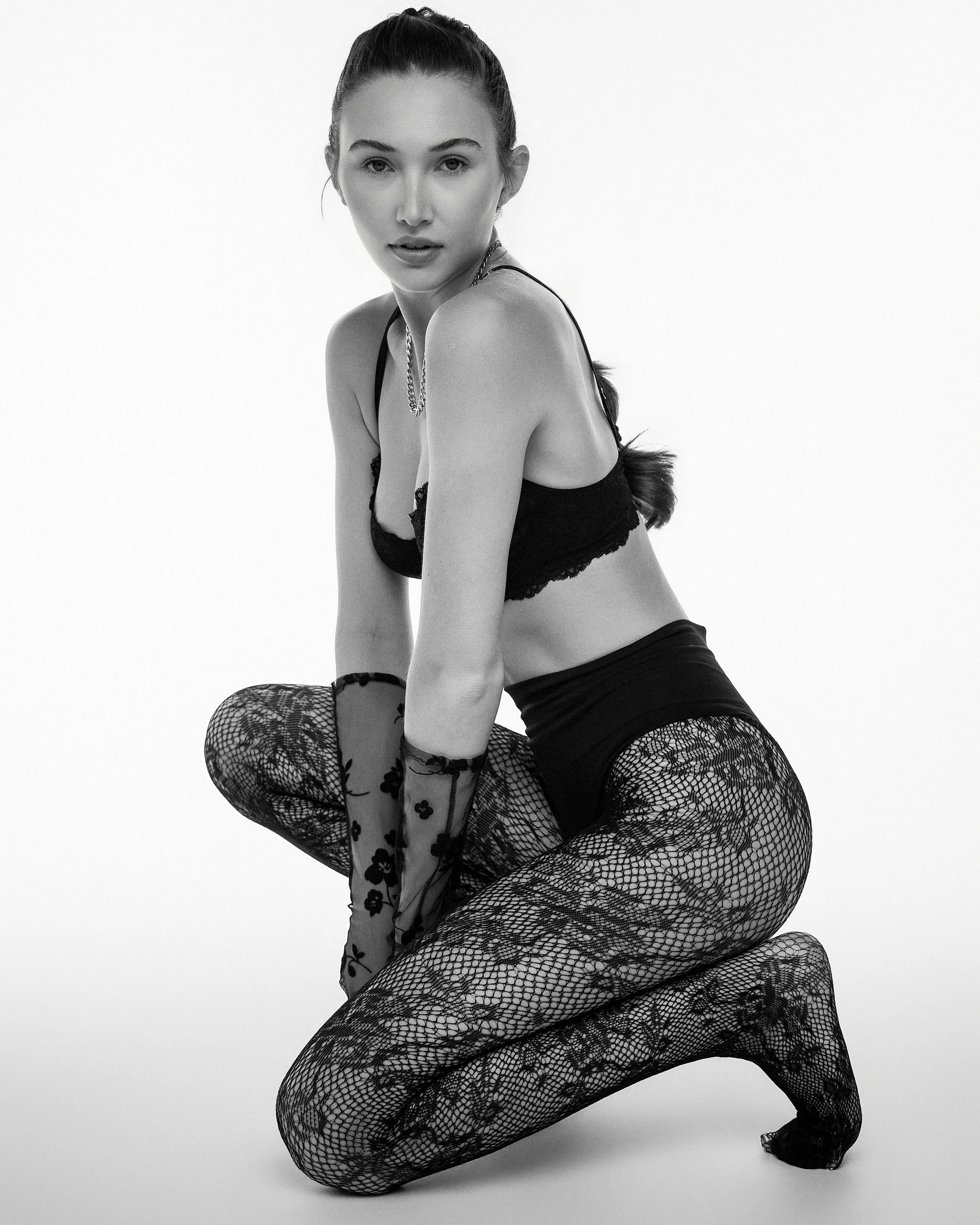Black and white photo of a woman in fishnet stockings, lace lingerie, and gloves, posing with curly hair and a serious expression.