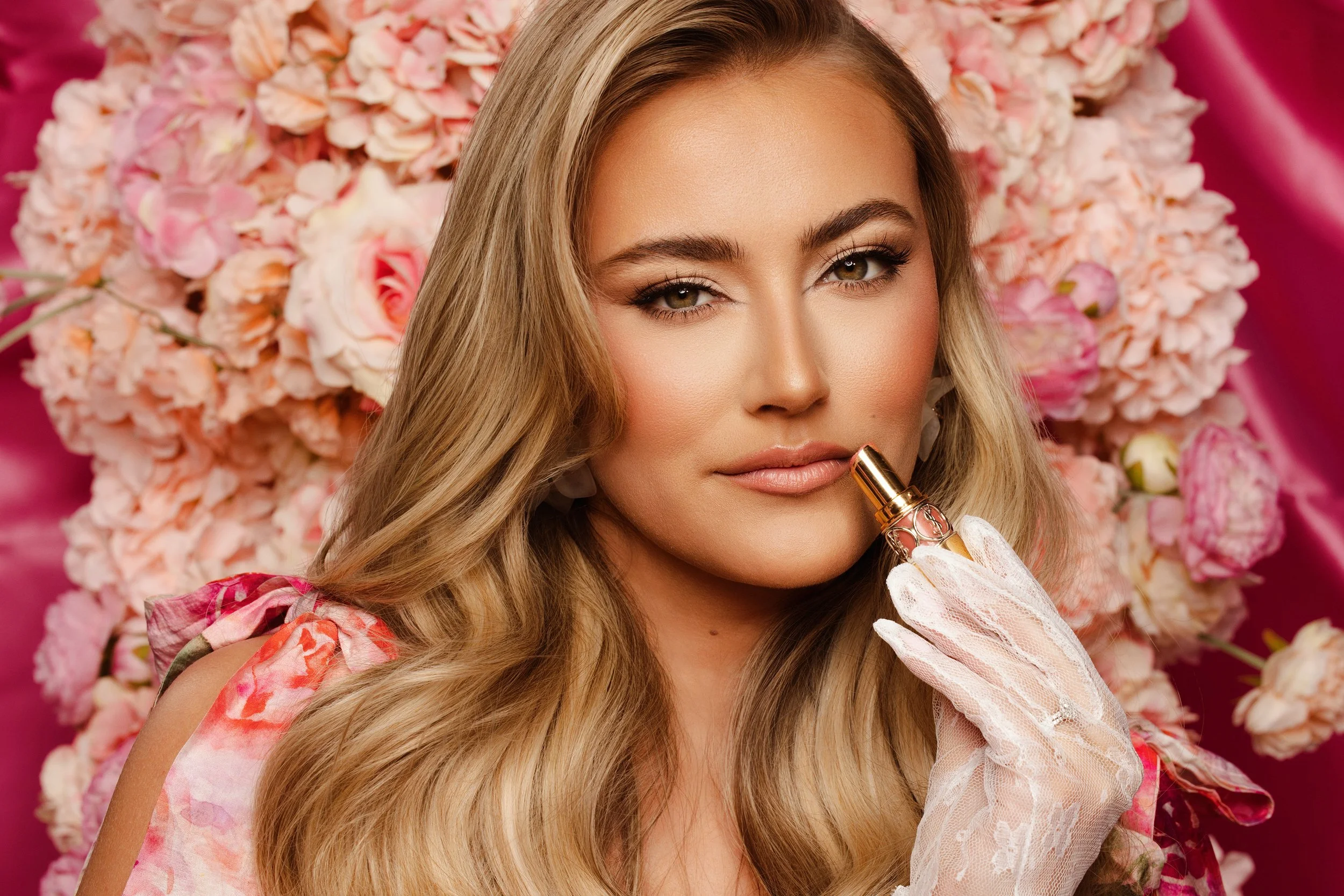 A woman with long blonde hair and makeup holding a lipstick near her lips, with a backdrop of pink and white flowers.