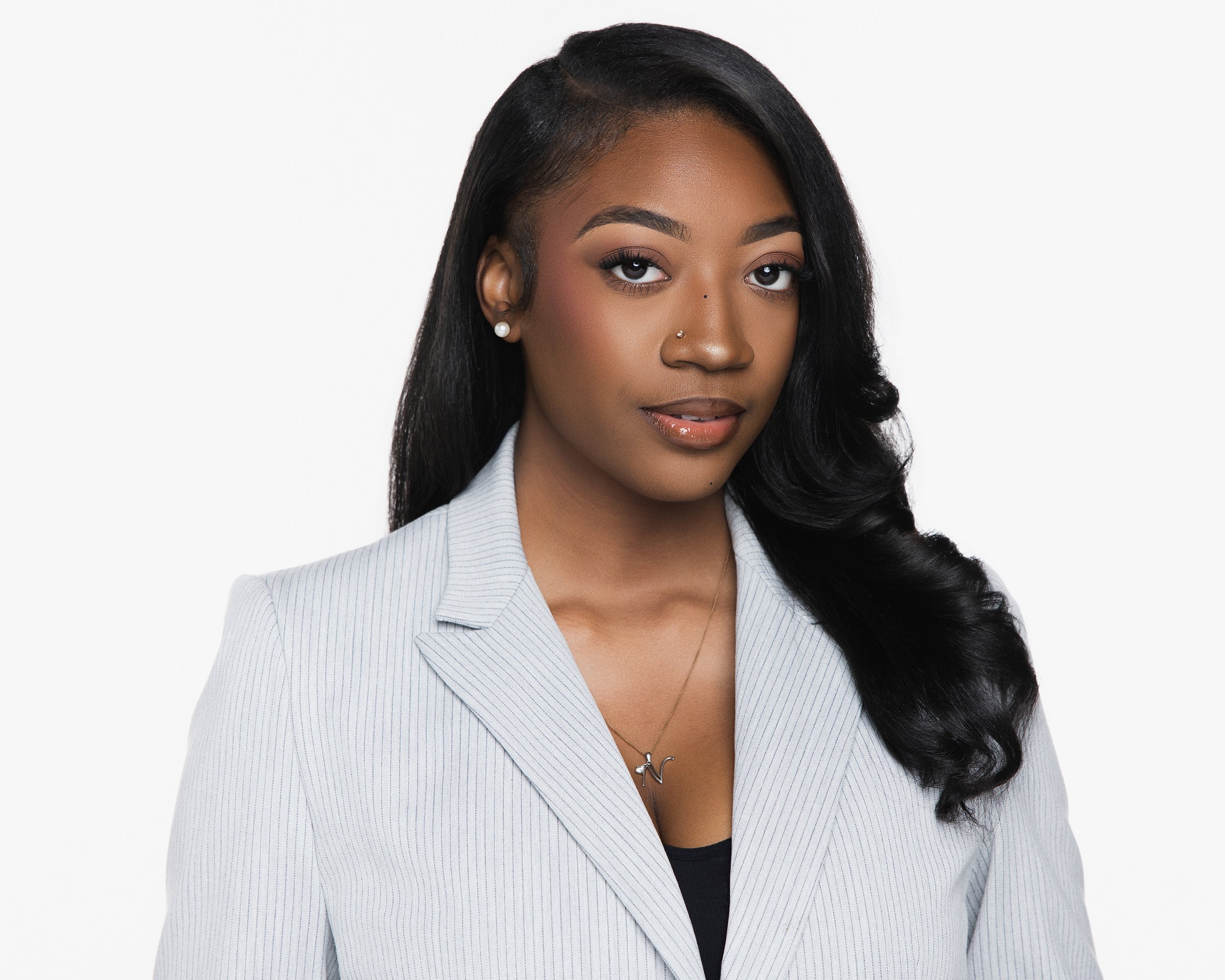 A young woman with long black hair, wearing a light gray striped blazer, pearl earrings, and a necklace with a pendant in the shape of a letter, posing against a white background.