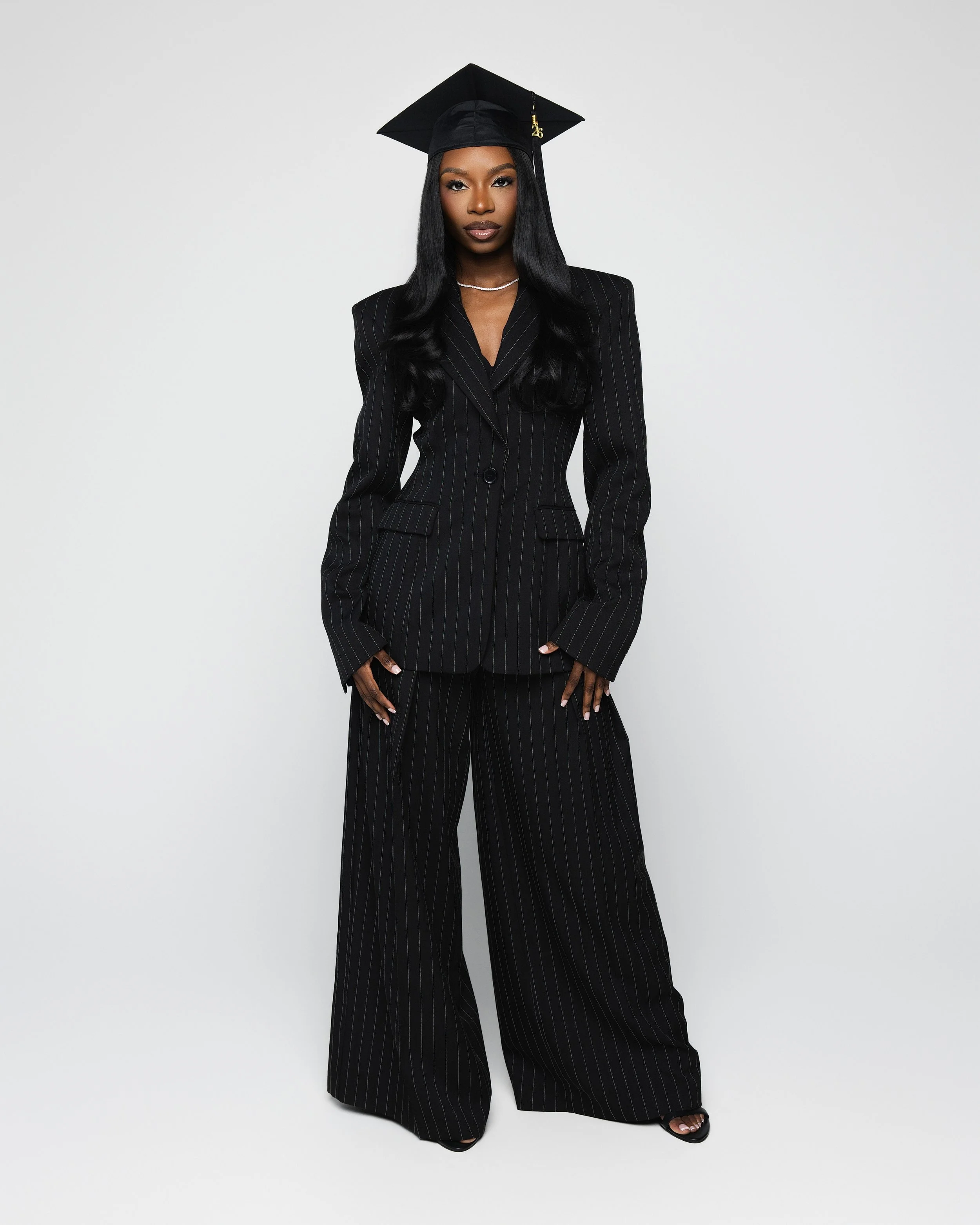 African American woman wearing a black pinstripe suit and mortarboard graduation cap, standing against a plain white background.