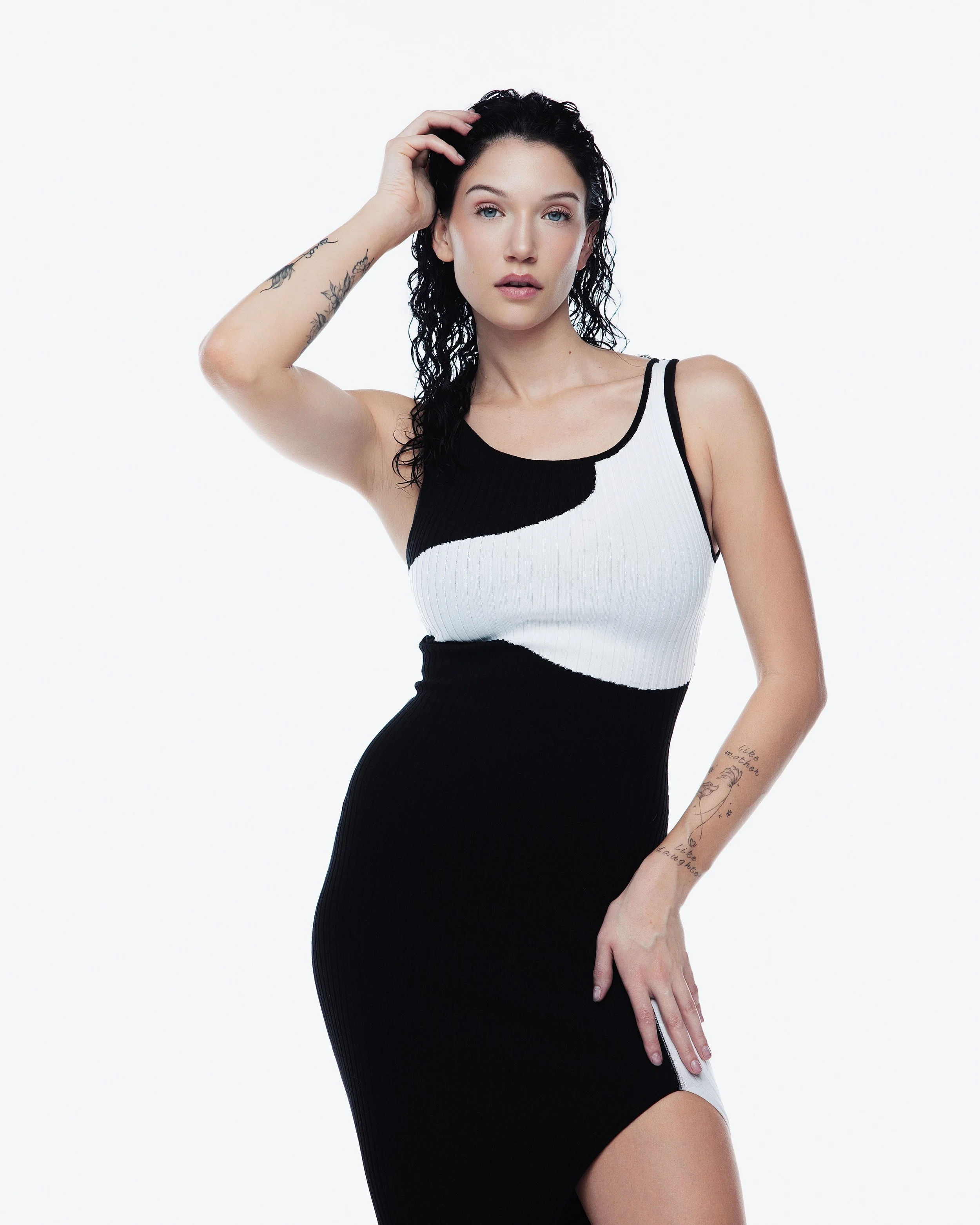 A woman with dark wet hair, light skin, and tattoos on her left arm, wearing a black and white sleeveless dress, posing against a white background.