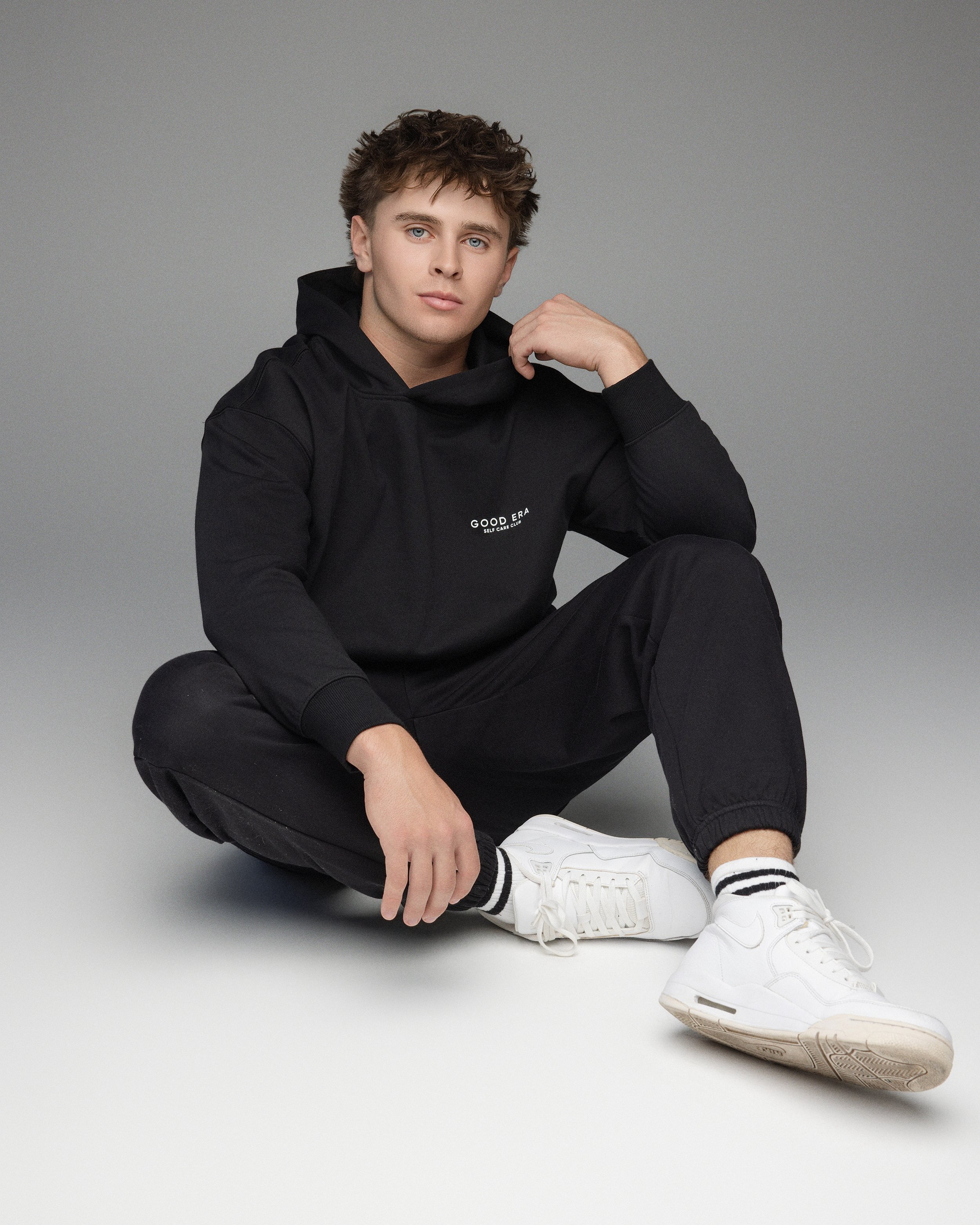 Young man sitting on the floor with one leg extended, wearing a black hoodie and black pants, white sneakers, and white socks with black stripes, against a plain gray background.