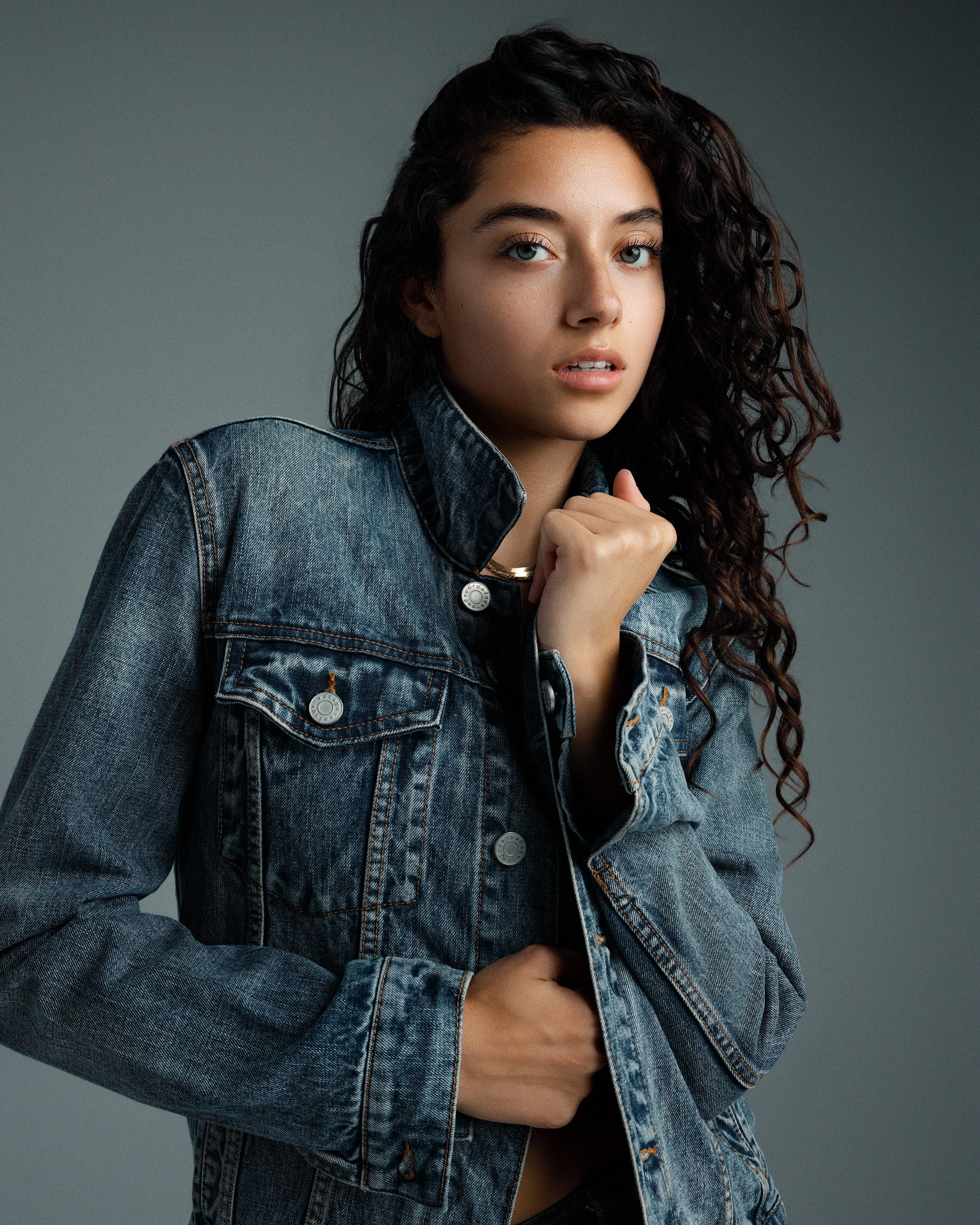 A young woman with curly dark hair and blue eyes wearing a denim jacket and a gold necklace. She is looking at the camera with a neutral expression against a gray background.