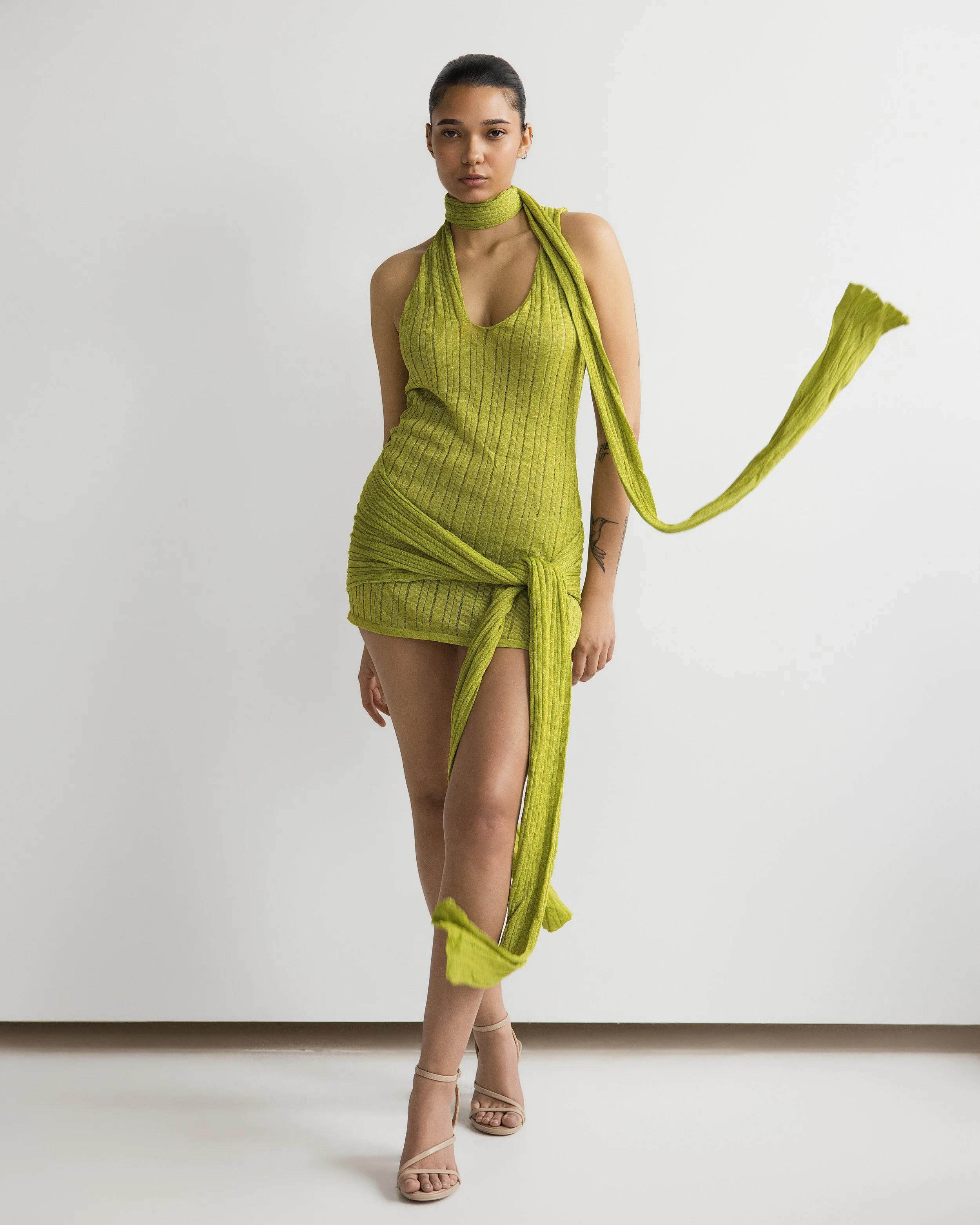 A woman wearing a bright green, sleeveless, ribbed dress with a matching scarf. She is standing against a plain white background, posing with one leg slightly forward and wearing beige high-heeled sandals.