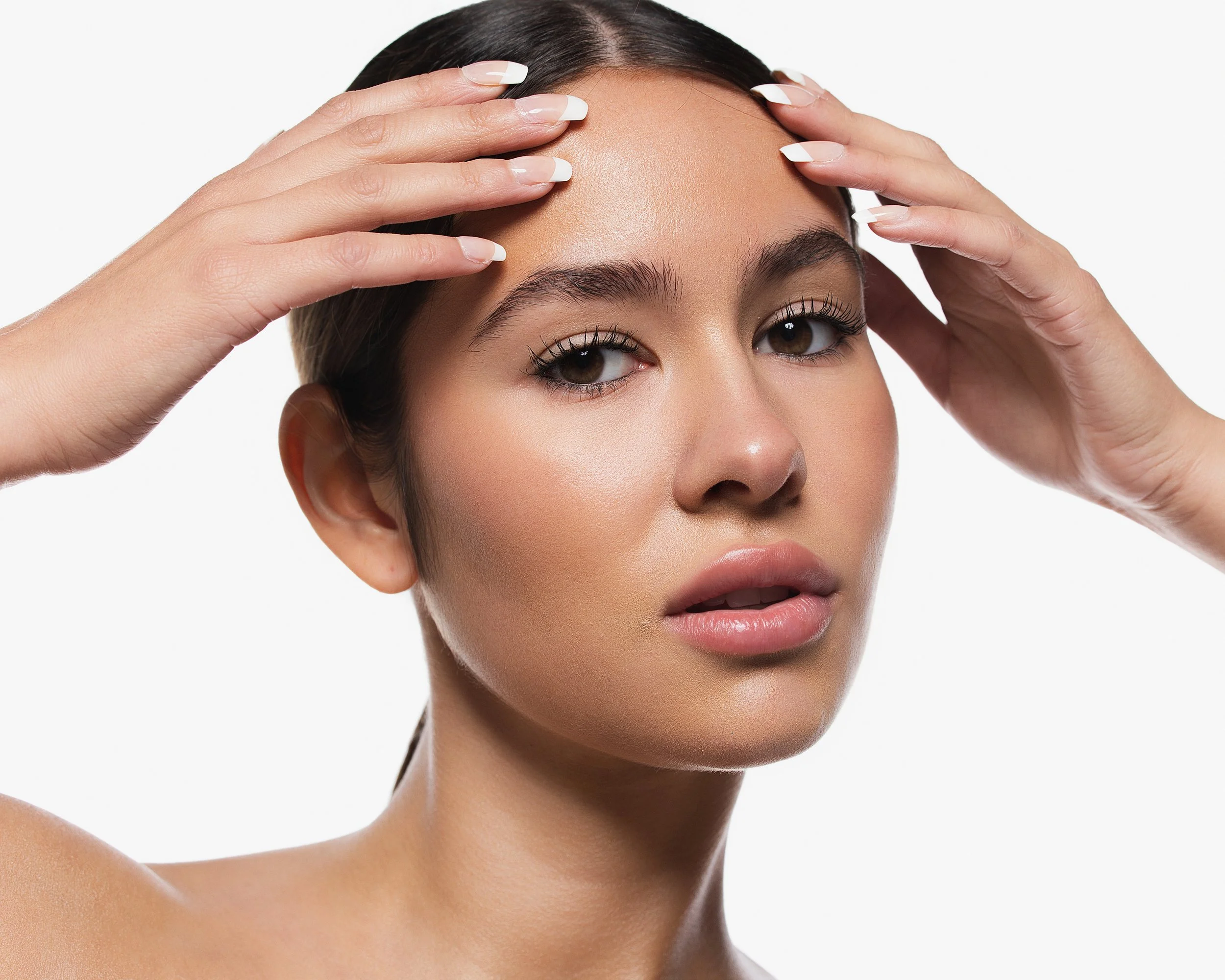 Close-up of a woman with dark hair, holding her forehead with both hands, with clean, white nails and natural makeup, set against a plain white background.
