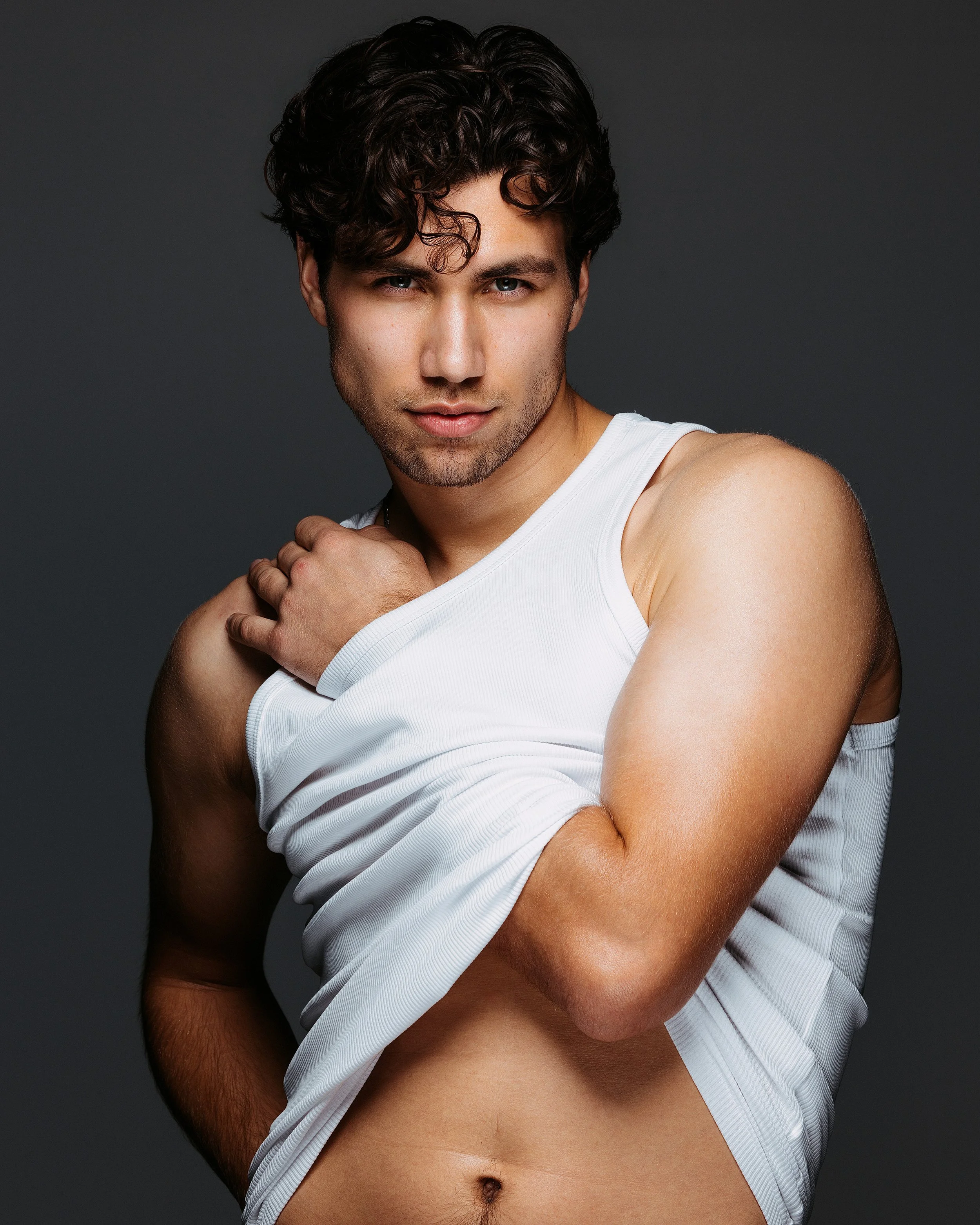 Close-up of a young man with dark curly hair, wearing a white sleeveless shirt, revealing his upper body and abdominal area, against a dark background.