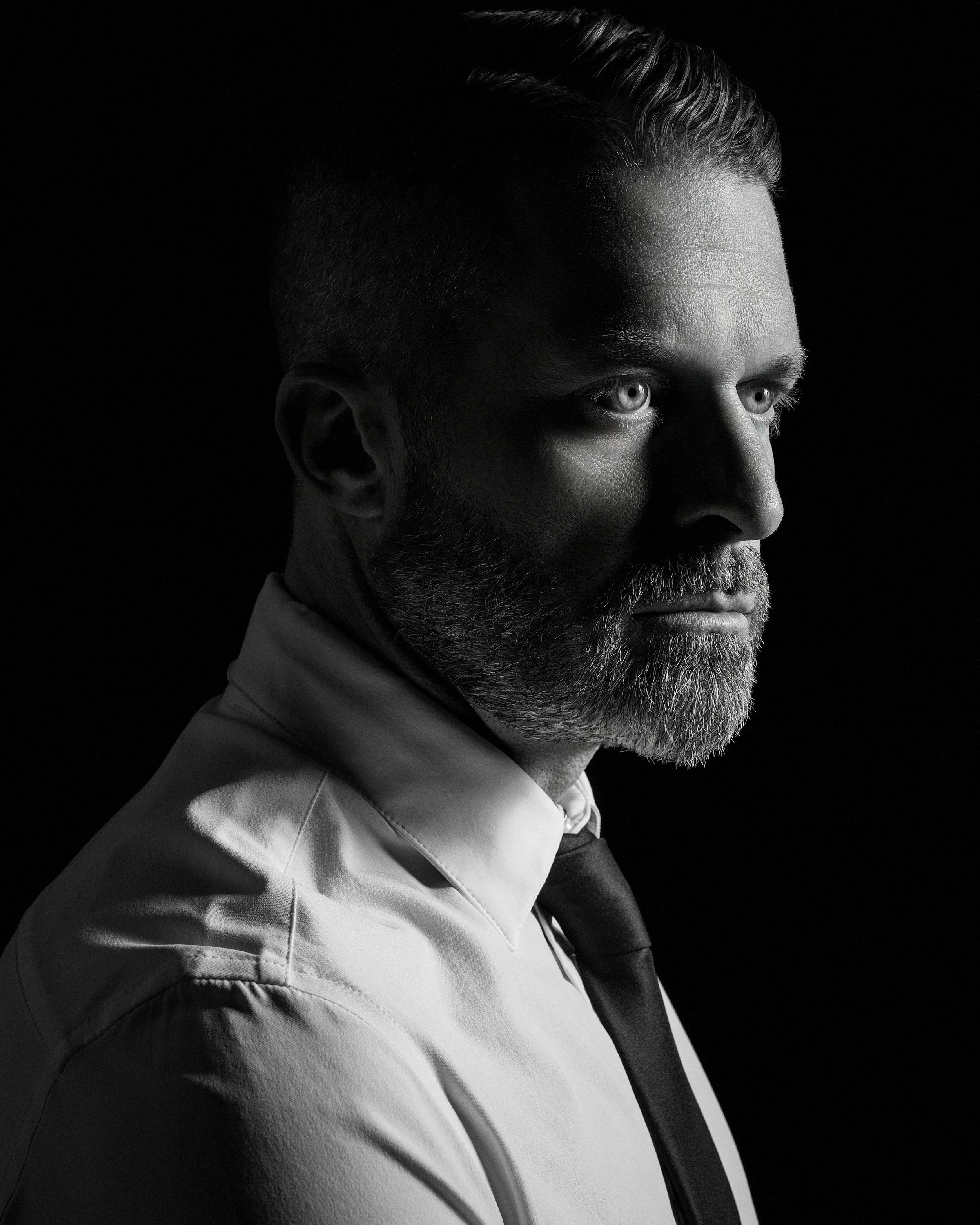 Side profile of a bearded man with slicked back hair wearing a light-colored jacket against a black background.