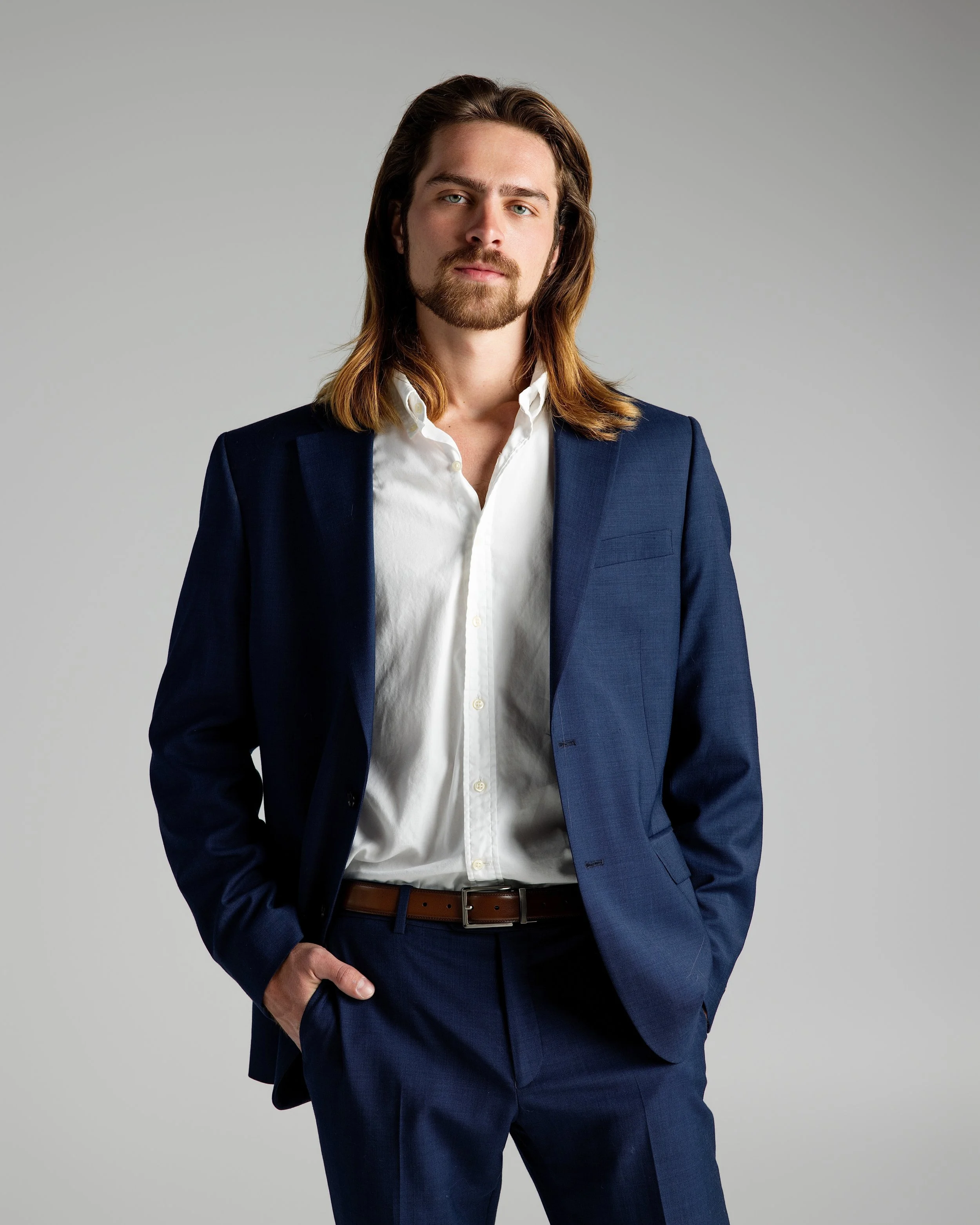 Portrait of a young man with long brown hair, light beard, wearing a navy blue suit, white shirt, standing against a plain grey background.
