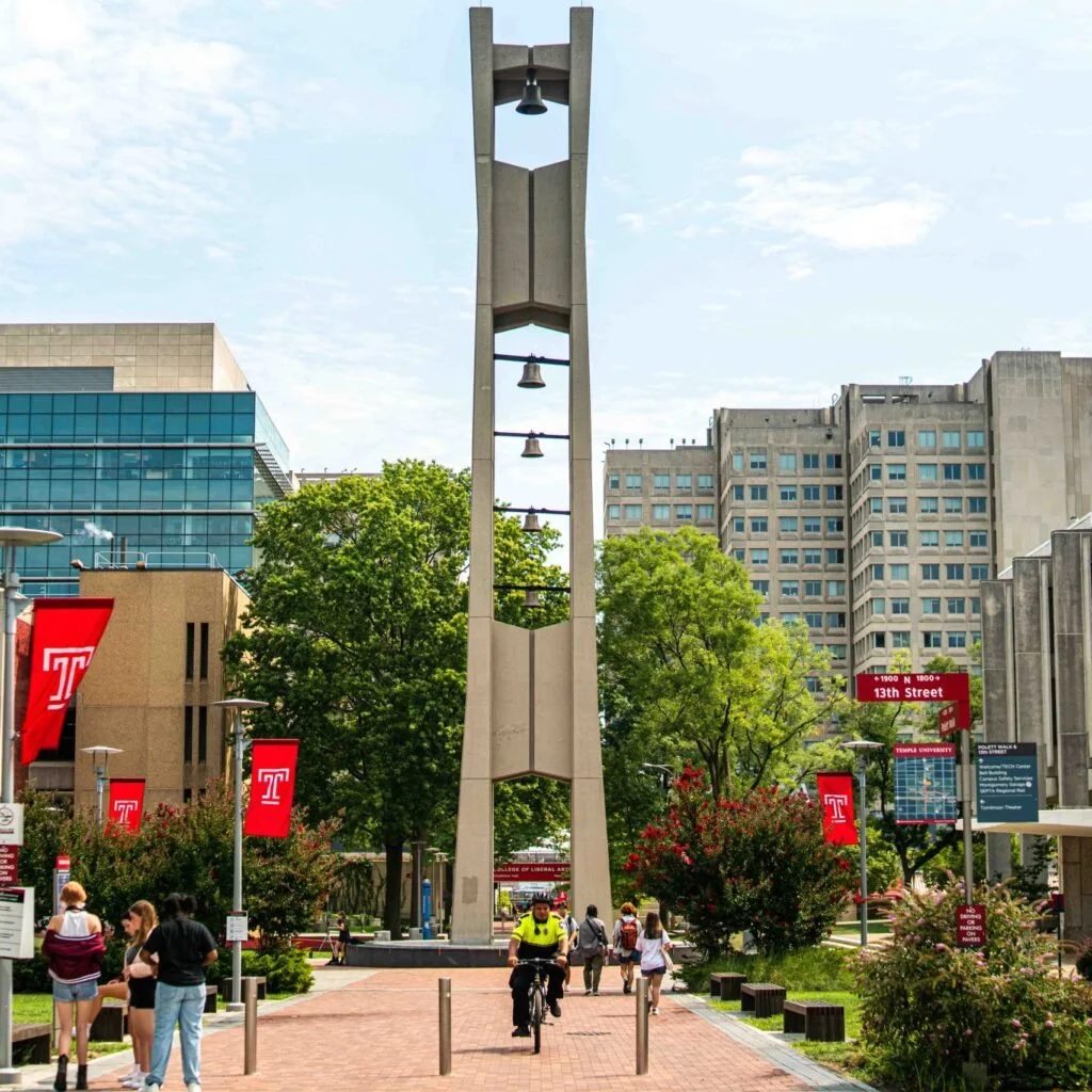 A university campus entrance with a tall bell tower in the center, surrounded by trees, people walking, and university banners, with modern buildings in the background.