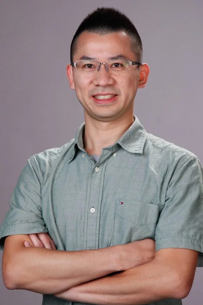 A man with glasses and short black hair, smiling with arms crossed, wearing a light gray button-up shirt, standing against a plain gray background.