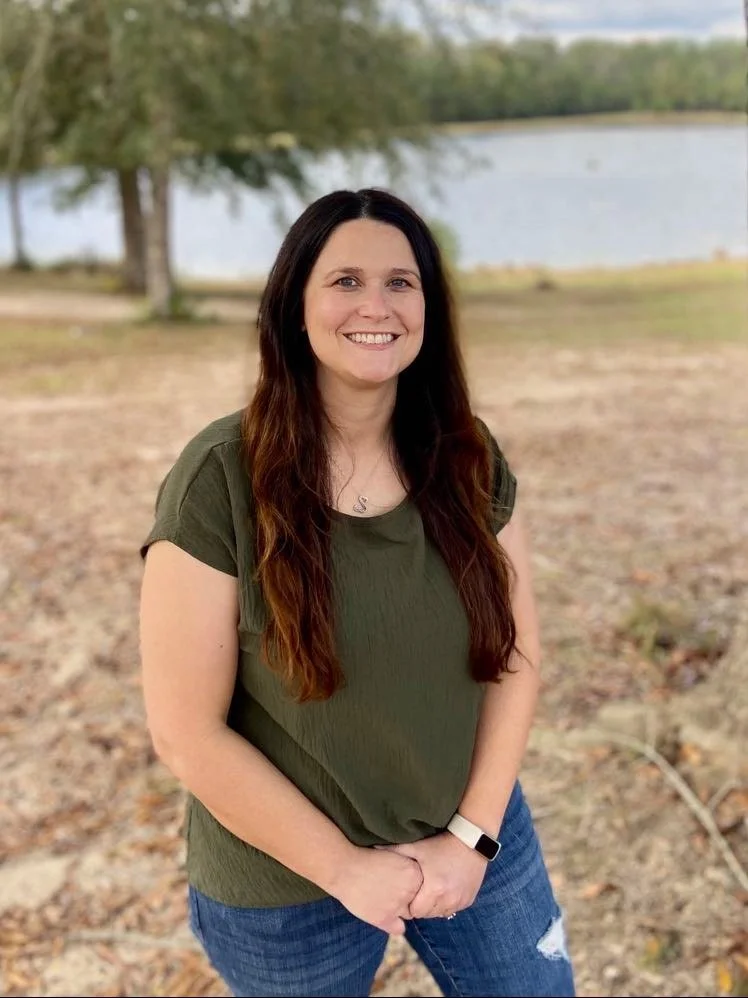 A woman with long brown hair wearing an olive green T-shirt, blue jeans, and a watch, standing outdoors near a lake with trees in the background, smiling at the camera.