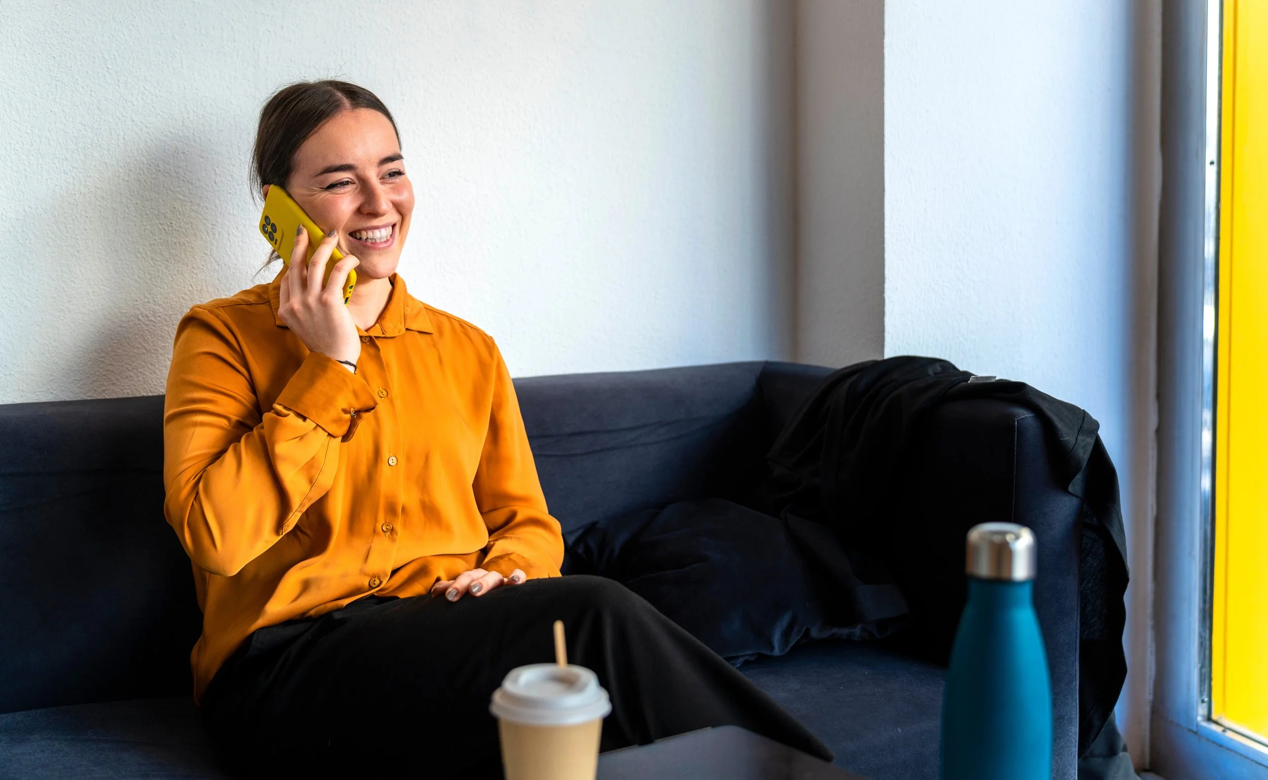 A young woman with brown hair in a ponytail, wearing an orange blouse, sitting on a black sofa, smiling and talking on a yellow cell phone. There is a coffee cup in front of her, and a backpack on the sofa beside her. She is near a window with sunlight and a yellow frame.