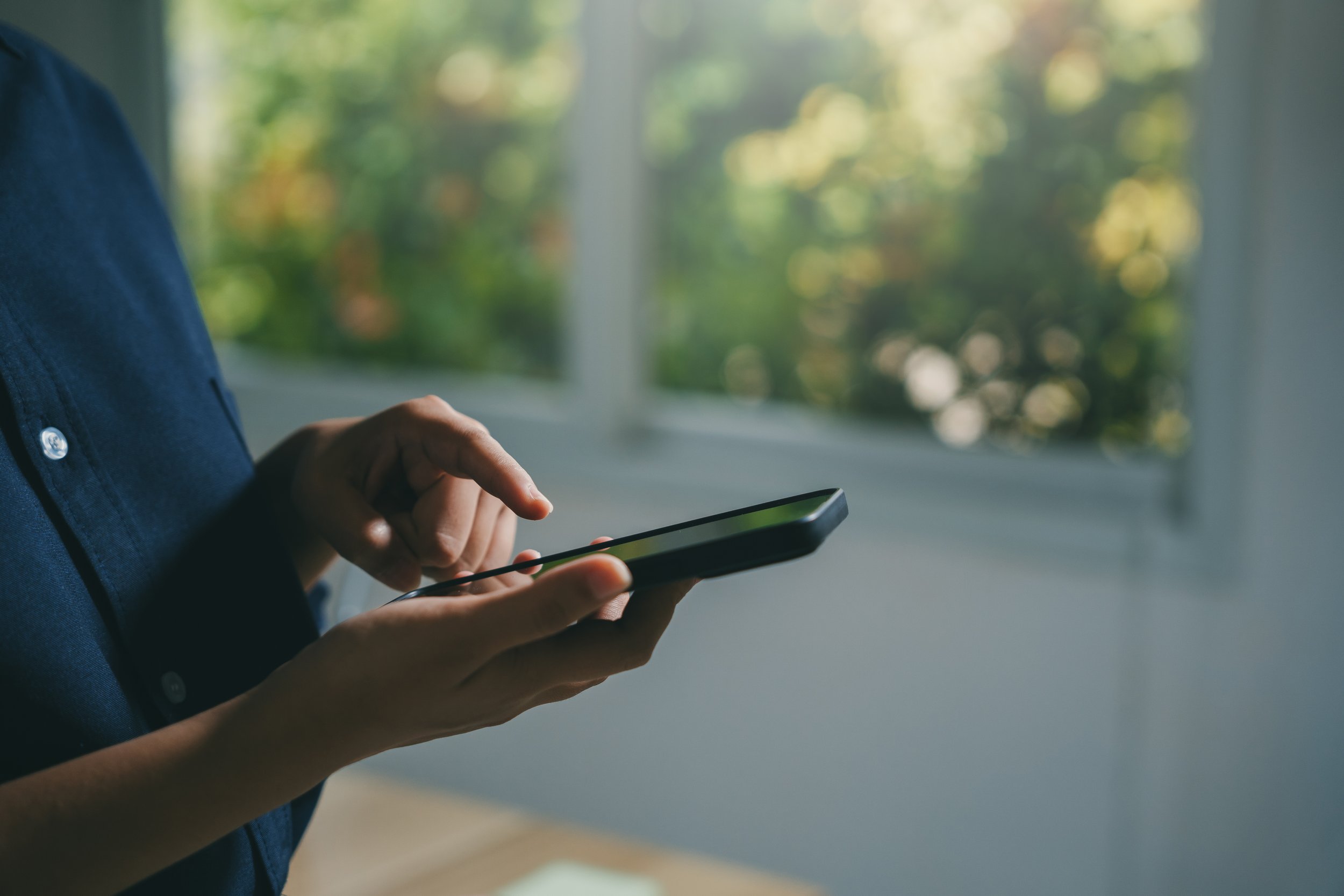 Person in a dark blue shirt using a smartphone, with a blurred window showing green trees outside in the background.