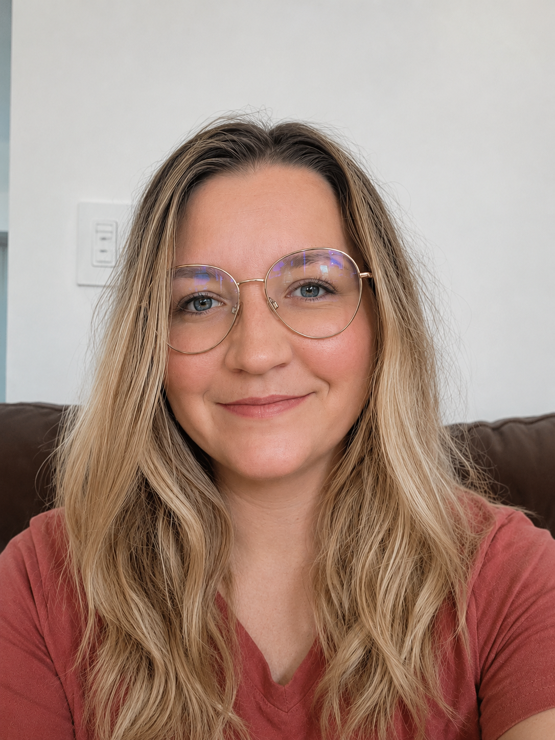 Close-up of a woman with long blonde wavy hair wearing round glasses and a reddish shirt, smiling while sitting on a brown couch in a living room with a white wall and a light switch.
