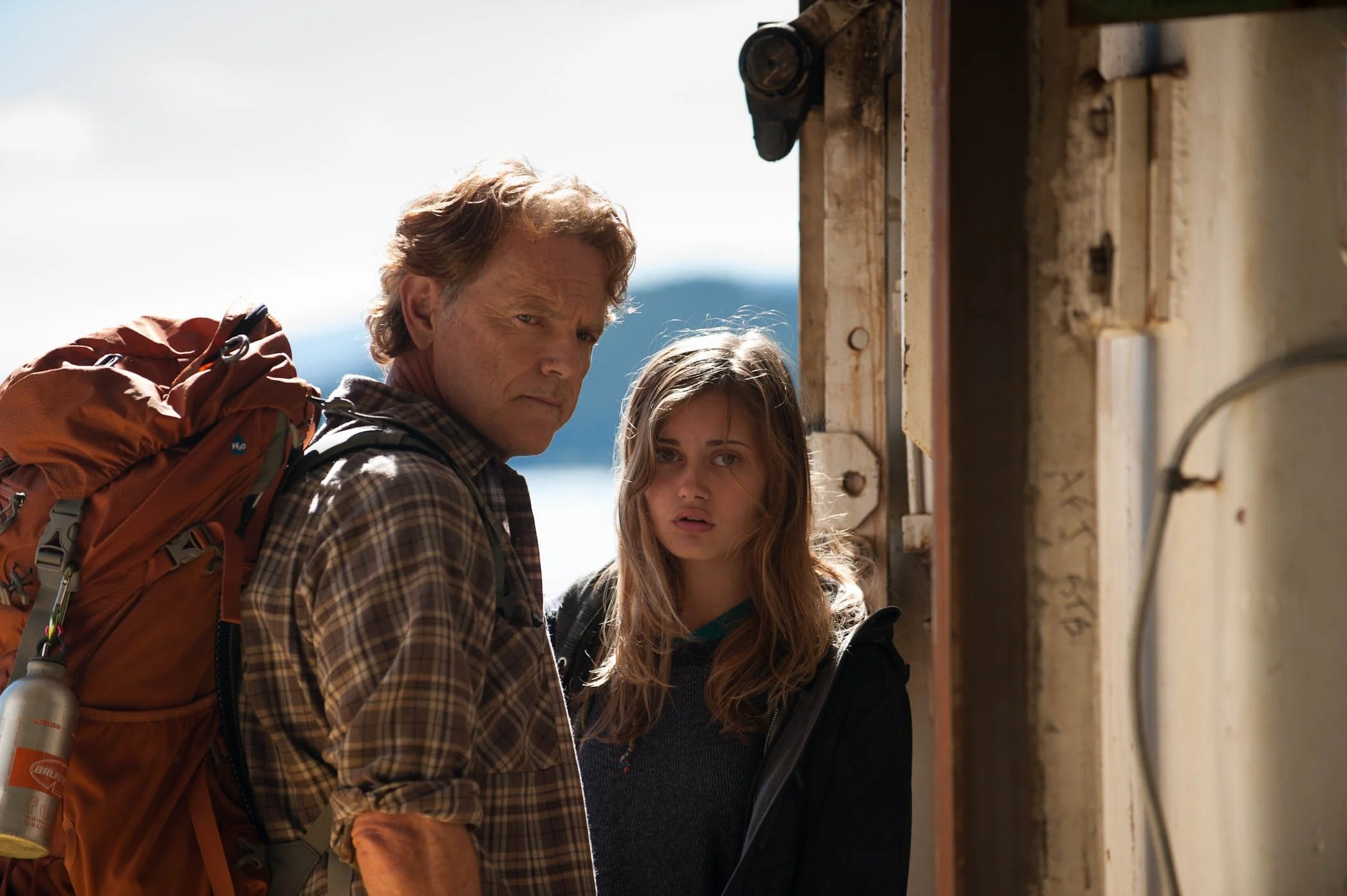 A man and a young girl looking at an electrical panel or circuit breaker box outdoors, with a mountain background and cloudy sky.