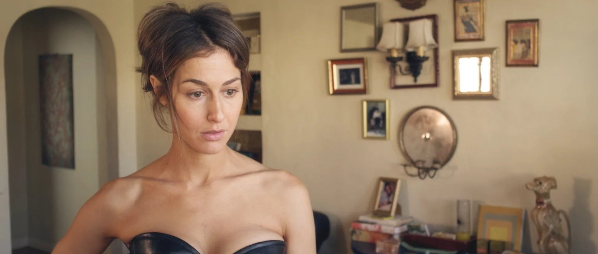 A woman with short brown hair looking down, wearing a strapless black top, in a room decorated with picture frames, lamps, and various decorative items on the walls and shelves.