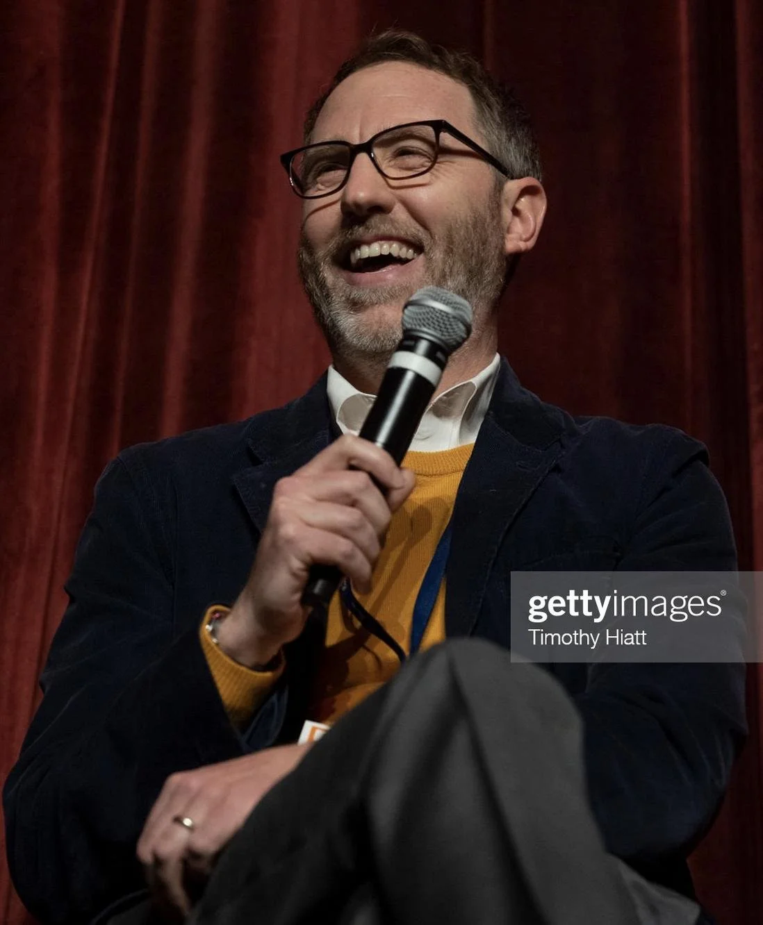 A man with glasses and a beard is smiling and talking into a microphone on stage with a red curtain background.