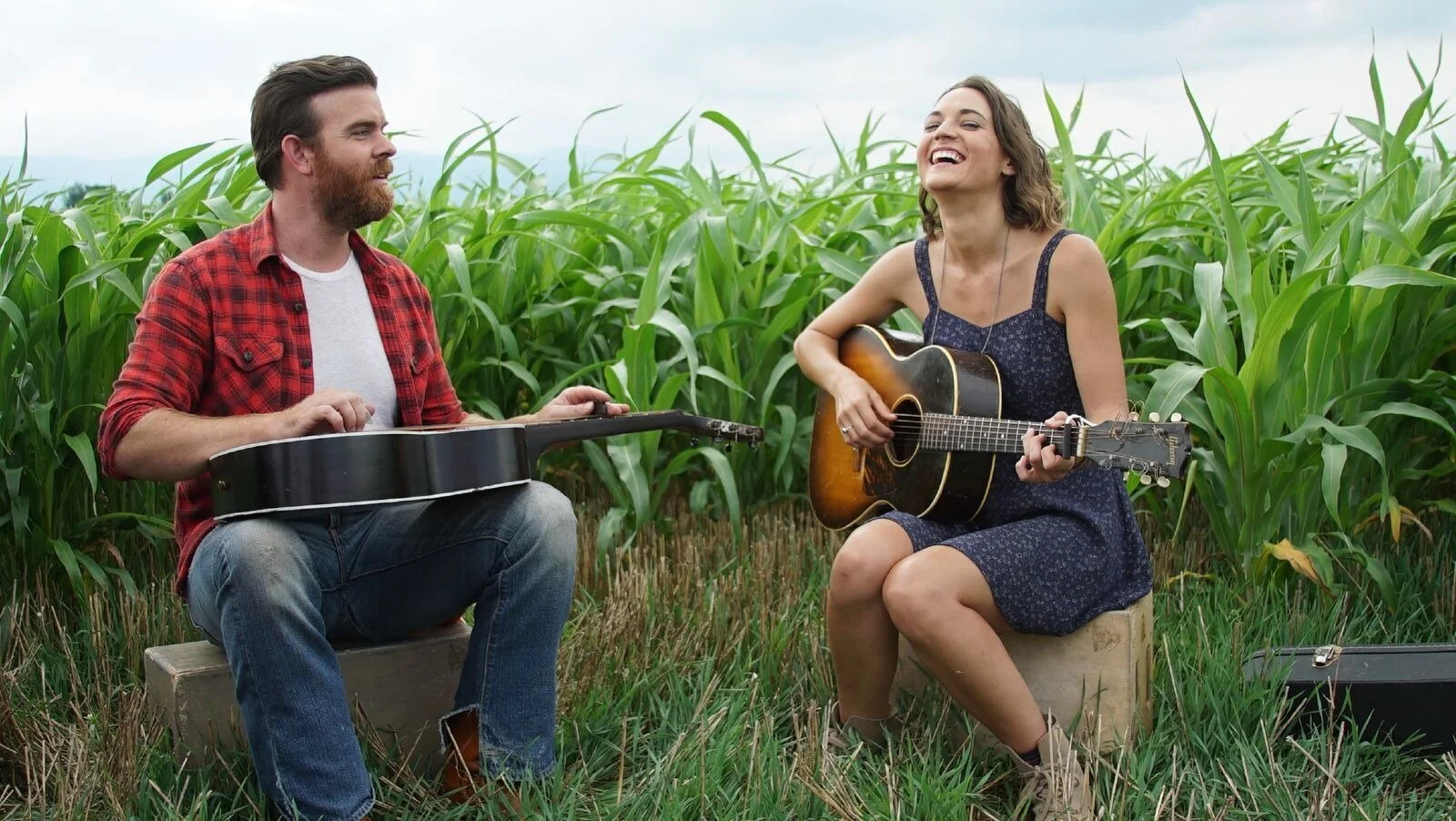 A man and a woman playing guitars and singing outdoors in a green field.