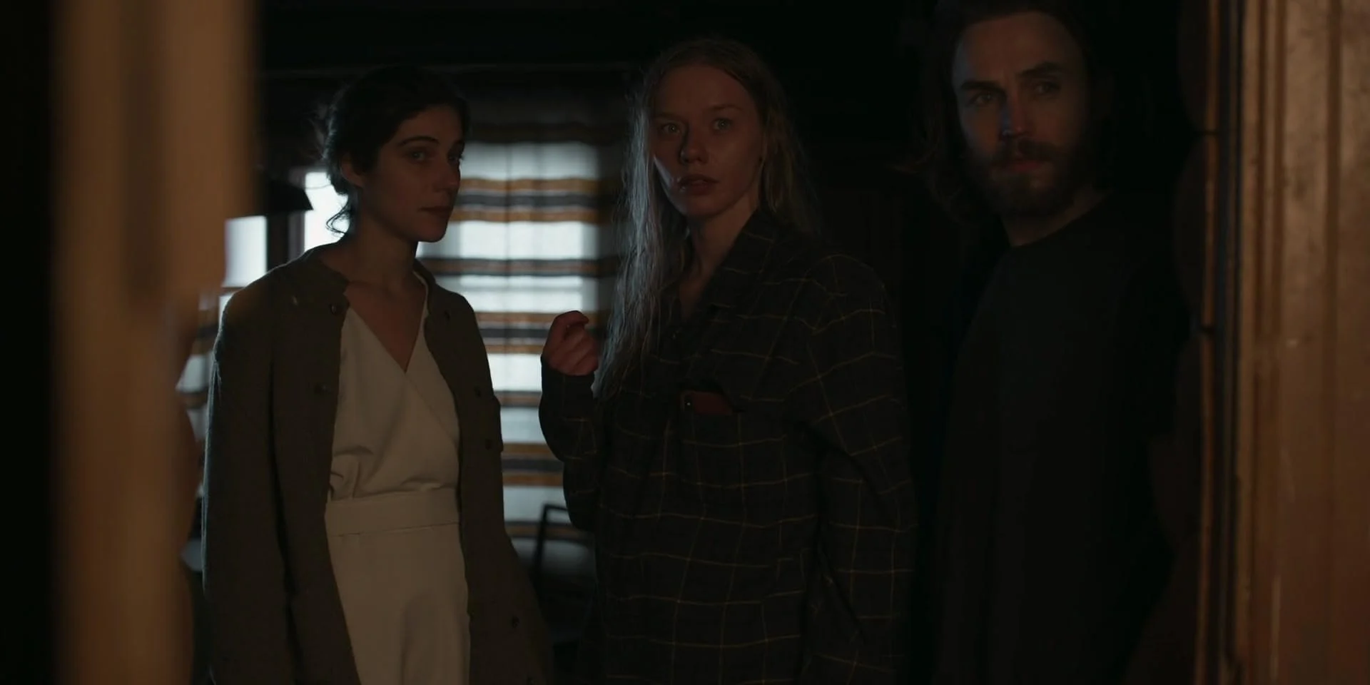 Three people standing in a dimly lit room, looking through a doorway with blinds behind them, with a serious or intense expression.