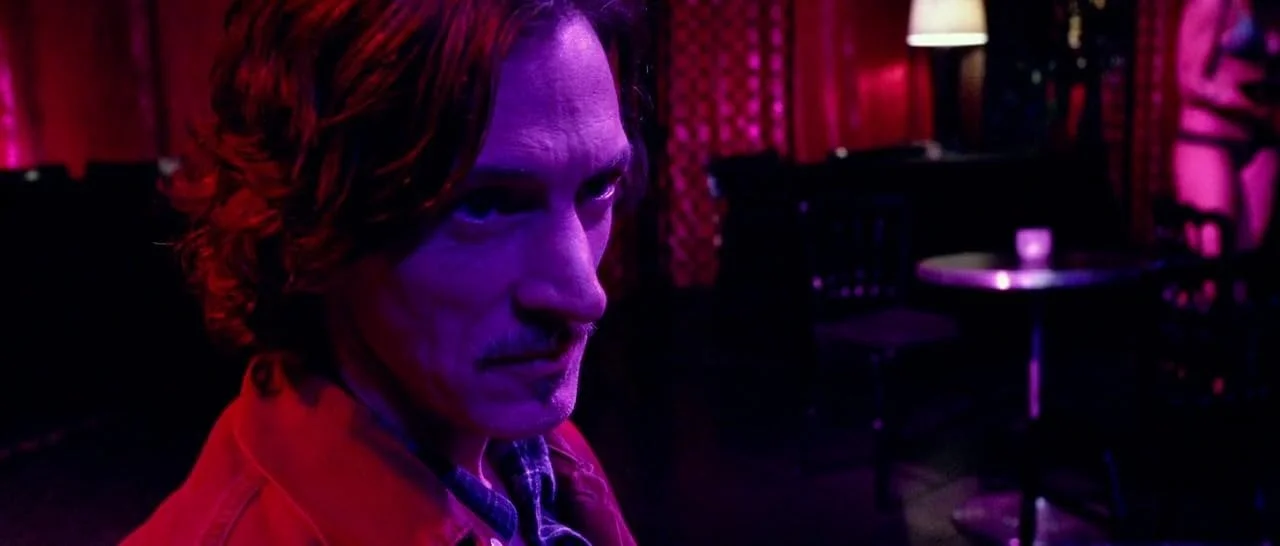 A man with wavy hair and a serious expression, wearing a red jacket, in a dimly lit room with red and purple lighting.