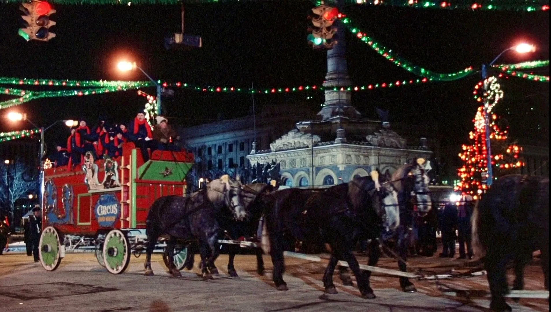 Night scene of a Christmas parade featuring a horse-drawn Santa Claus float decorated with red and green colors, children dressed as elves riding the float, and festive string lights overhead. In the background, there is a historic building and a str