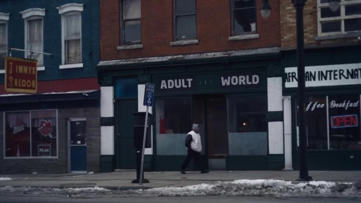 A man walking past a nightclub with a sign that reads 'Adult World', with snow on the sidewalk and a neon 'Open' sign in a window.