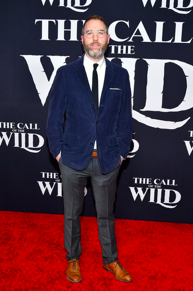 Man with glasses and beard in blue blazer, white shirt, black tie, gray pants, and brown shoes standing on a red carpet in front of a dark backdrop with text for the "Call of the Wild" movie.