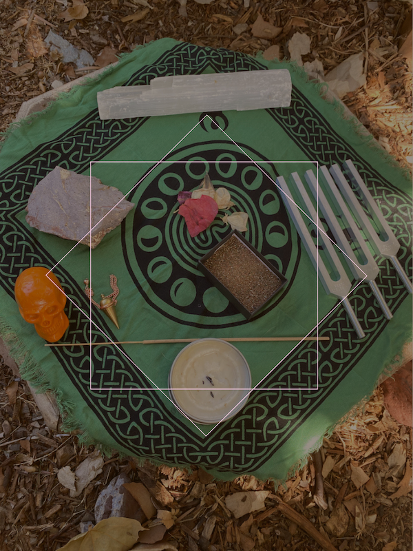 An outdoor altar with various spiritual objects on a green cloth with black designs, surrounded by fallen leaves.