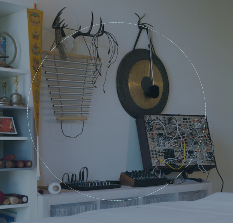 A collection of musical and electronic items on a shelf, including a handcrafted percussion instrument made of wooden sticks, antler decor, a gong, and a synthesizer with exposed wiring.