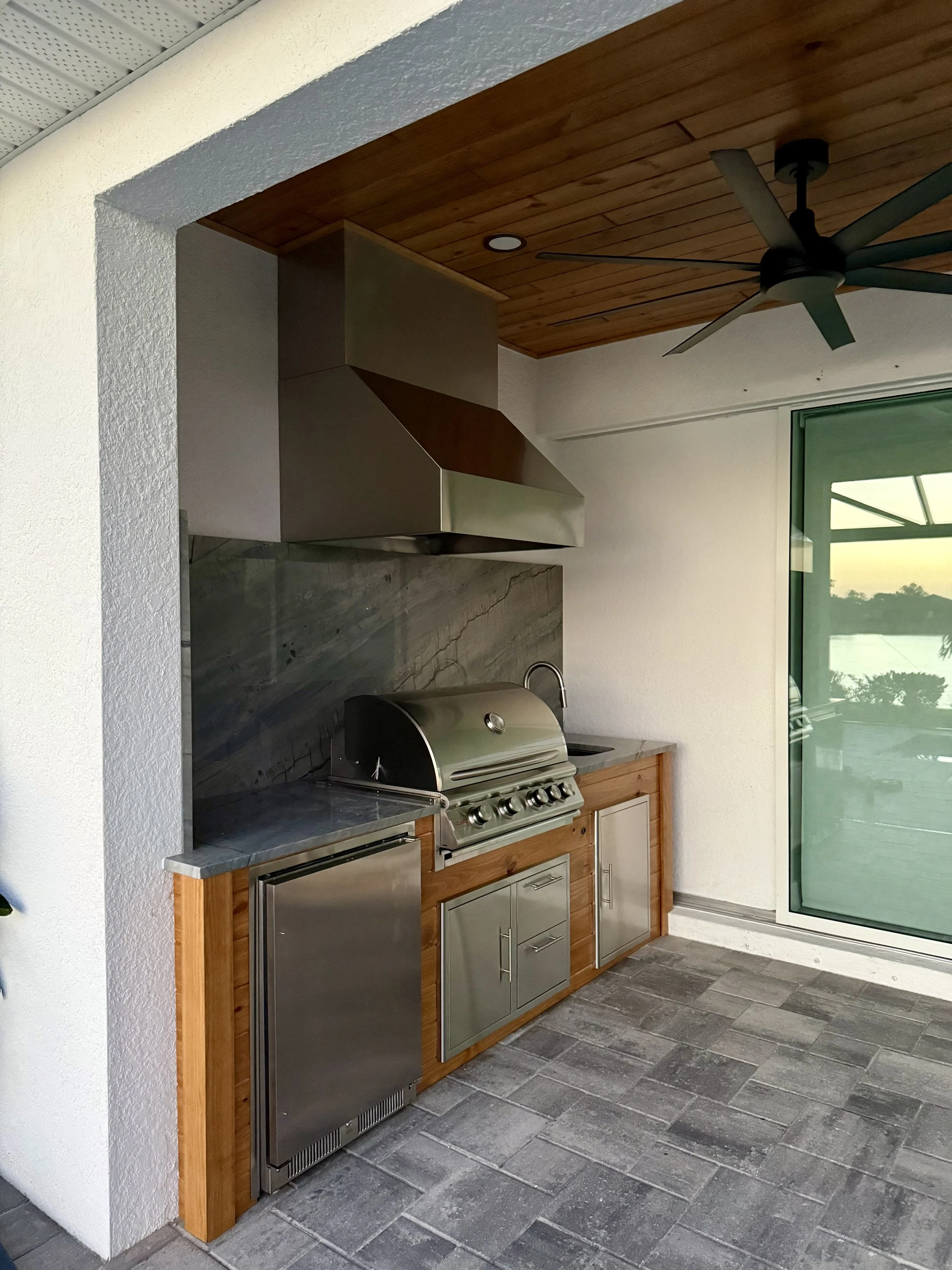 Outdoor kitchen area with stainless steel grill, wooden cabinets, marble countertop, ceiling fan, and large glass sliding door with a view of a body of water and trees outside.