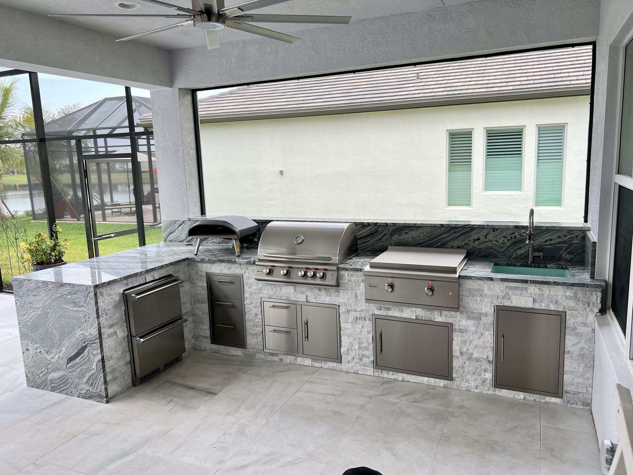 Outdoor kitchen with a built-in grill and marble countertops, enclosed by screened walls showing a backyard with a pond, patio furniture, and trees.