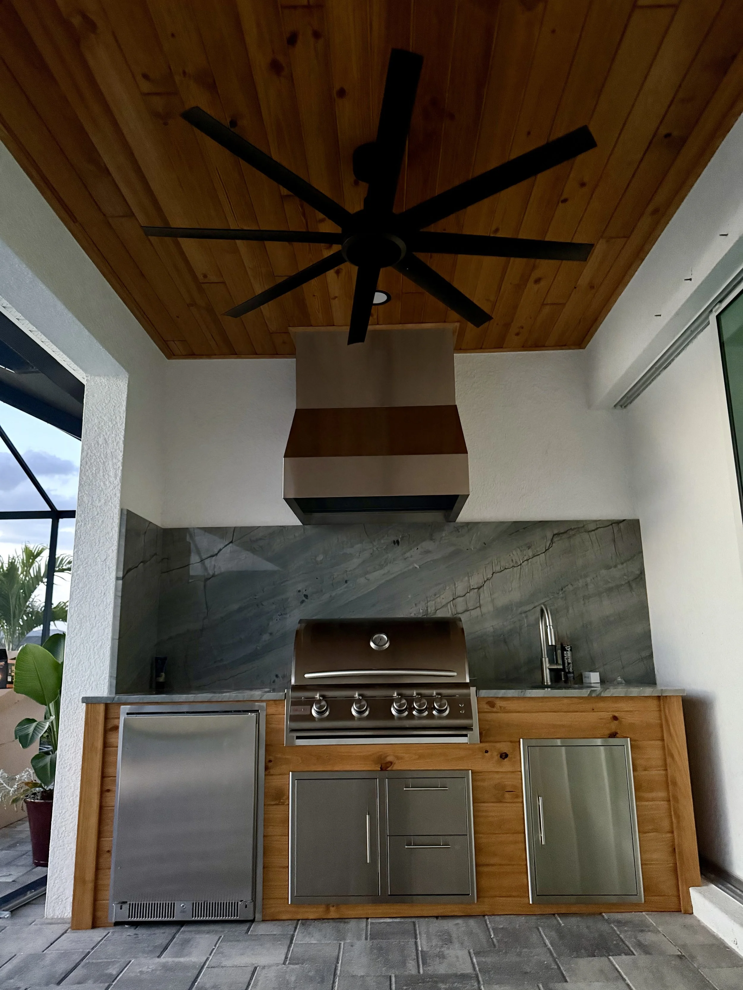 Outdoor kitchen with wooden cabinets, stainless steel appliances, grill, and a vent hood, under a wooden ceiling with a ceiling fan.