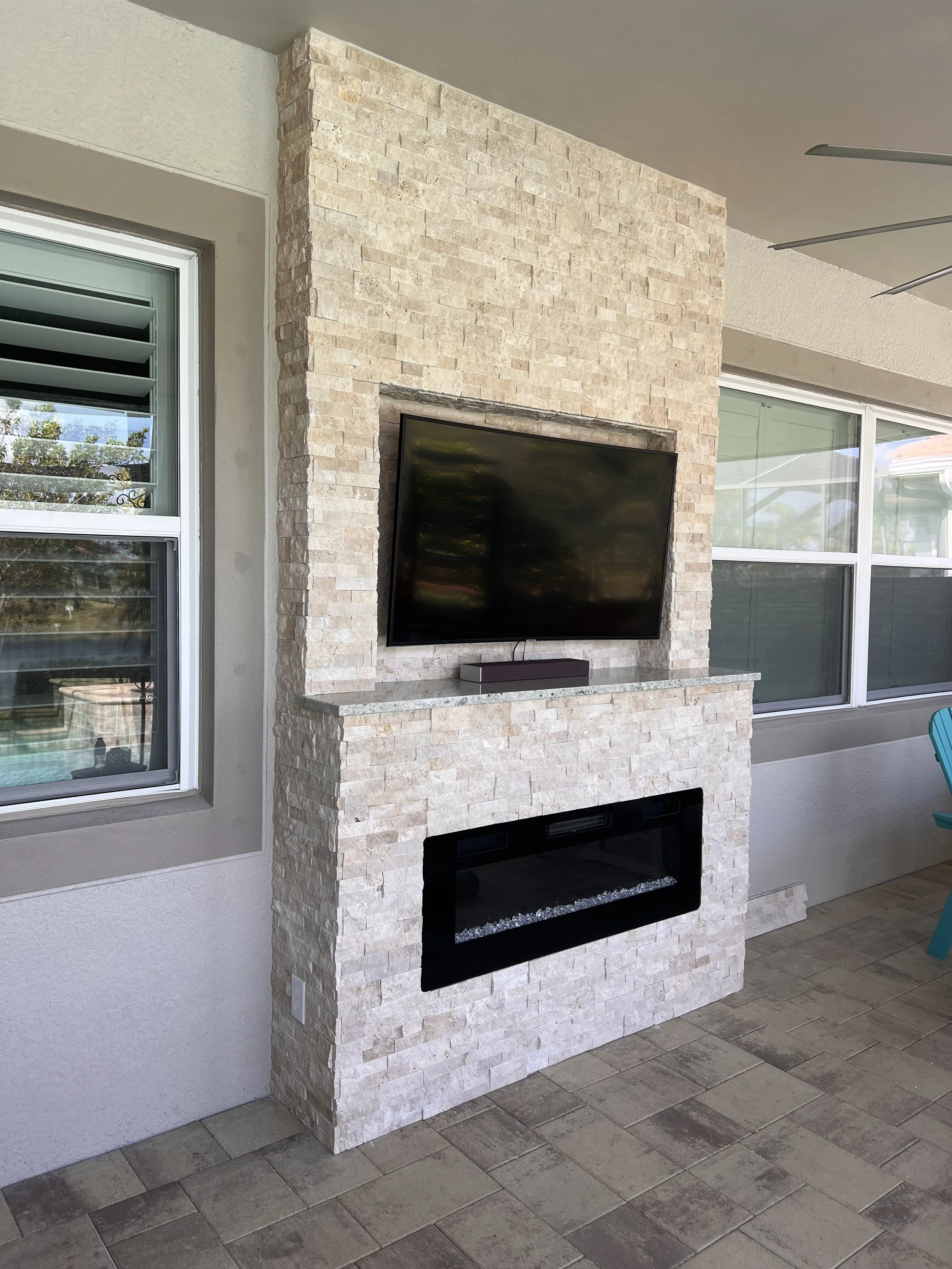 Outdoor fireplace with a beige stone surround, mounted TV, and large window to the left. Floor is tiled with grey stones.