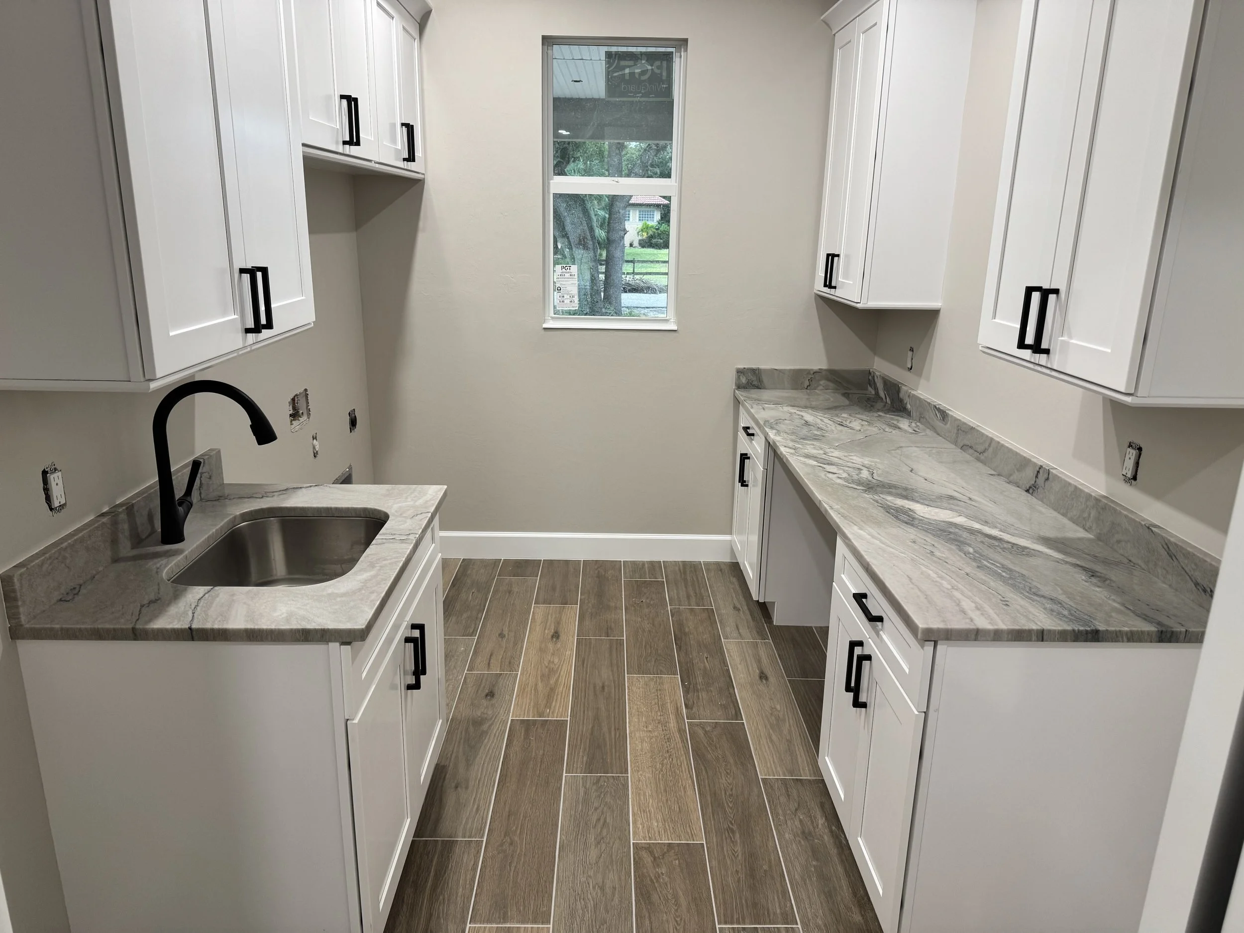 Empty kitchen with white cabinets, gray and white marble countertops, a black kitchen faucet, and a window overlooking trees and a house outside.