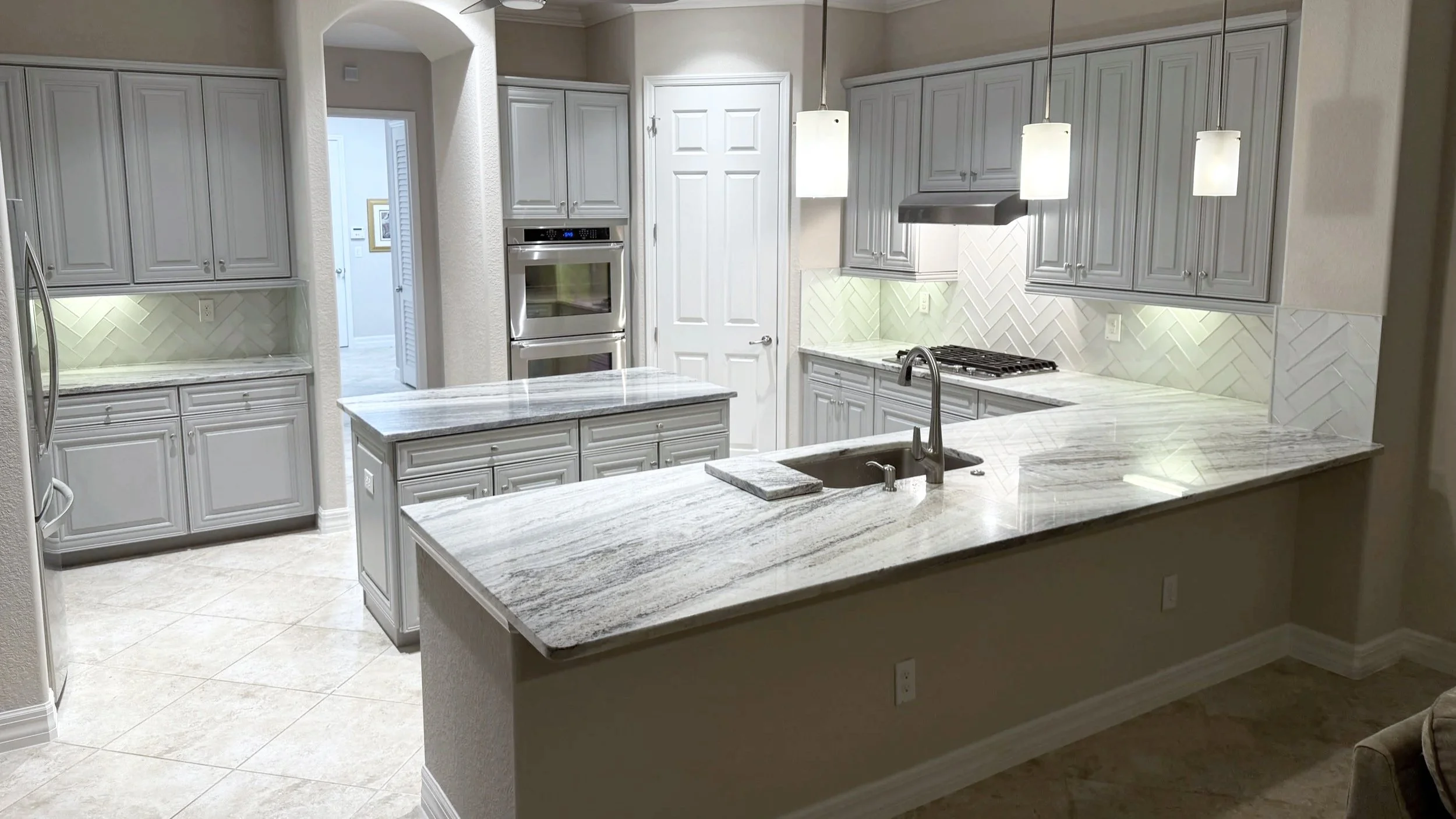 A modern kitchen with light gray cabinets, white and gray marble countertops, a center island, a double oven, a stainless steel refrigerator, pendant lighting, and a herringbone tile backsplash.
