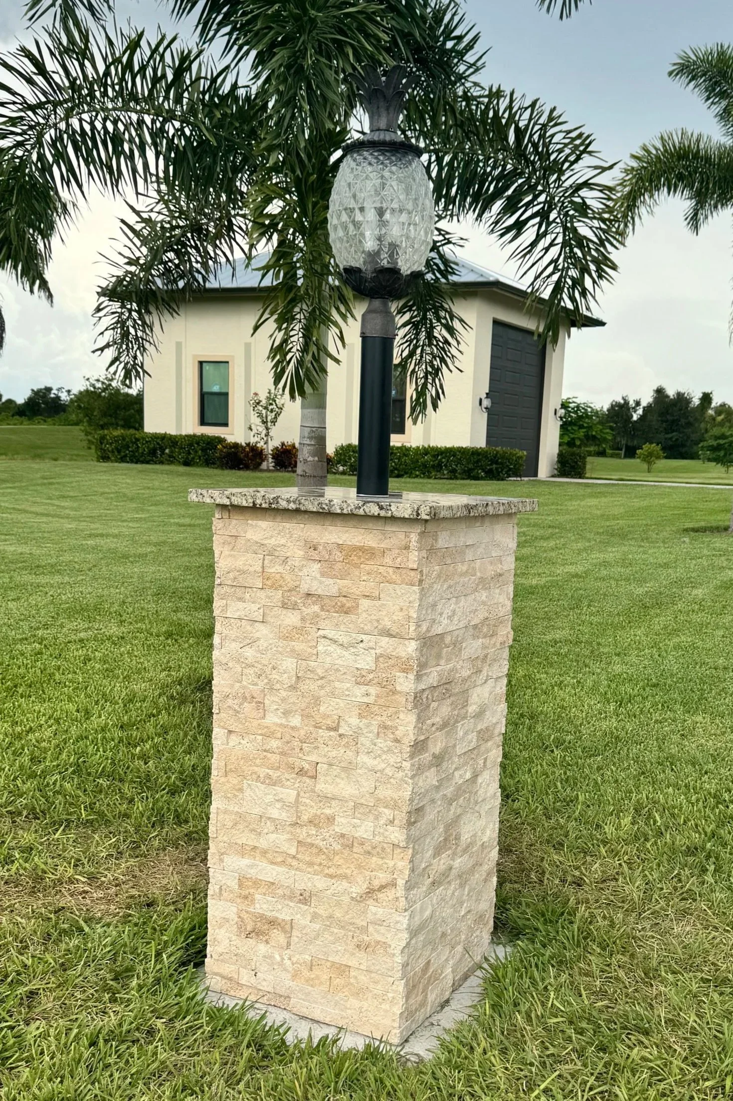 A lamp post with a glass lantern is situated on a stone pedestal in a grassy yard, with a house and trees in the background.
