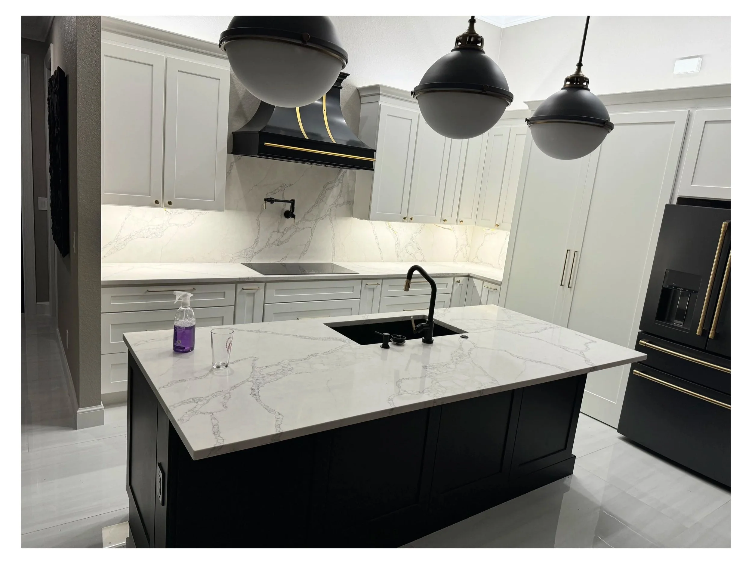 Modern kitchen with white cabinetry, black accents, a marble island, and three black pendant lights hanging from the ceiling.